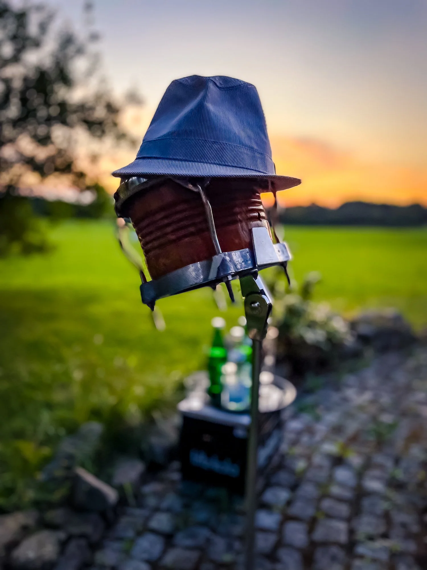 Nahaufnahme einer Trommel eines Schlagzeugs, auf der ein Hut liegt. Im Hintergrund eine Wiese, ein Baum und ein Sonnenuntergang.