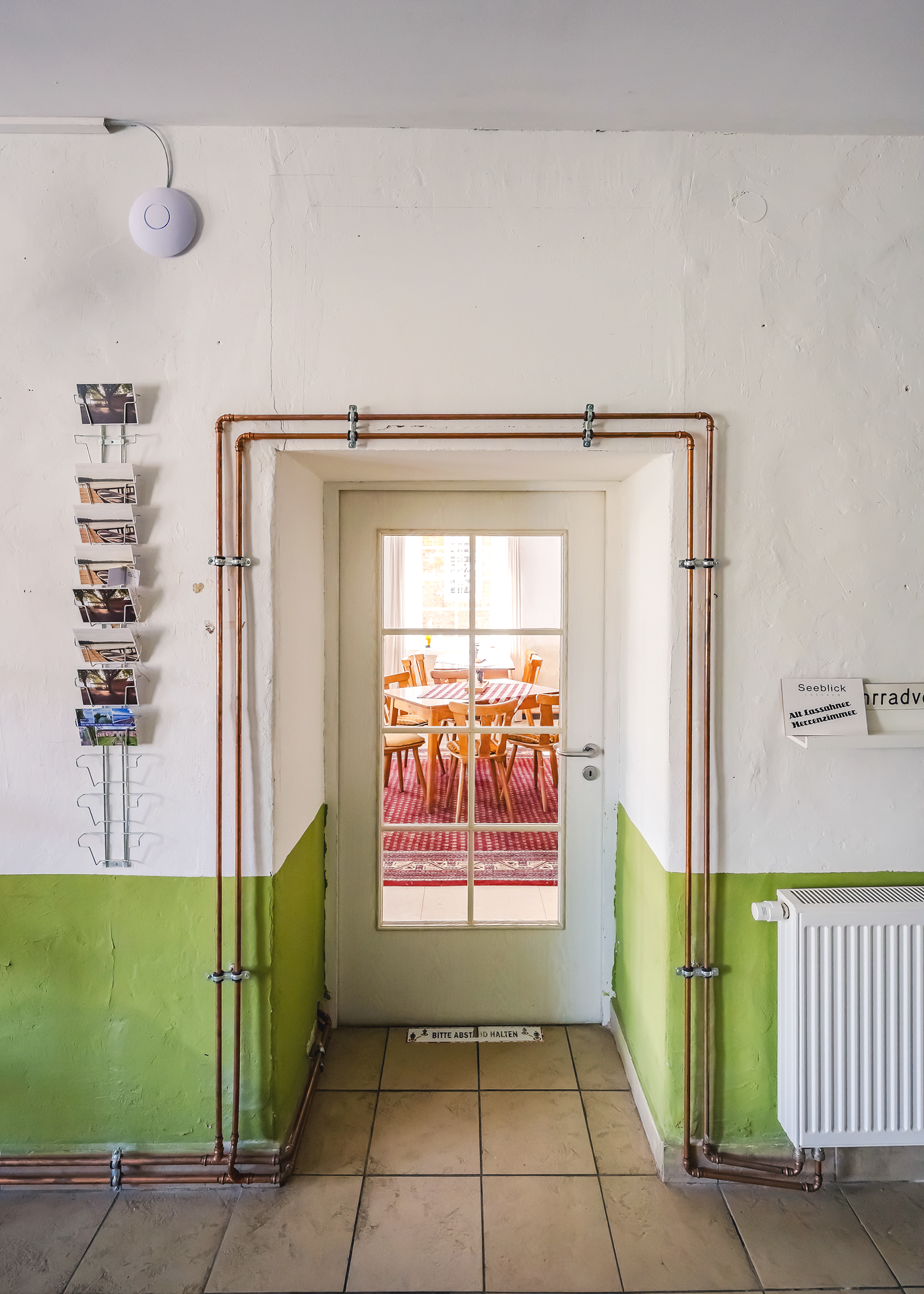 Ein Eingangsraum mit weißen Wänden, bepflanzten Kupferrohren und einer gläsernen Tür, die in einen hell erleuchteten Raum mit einem runden Holztisch und Stühlen führt.