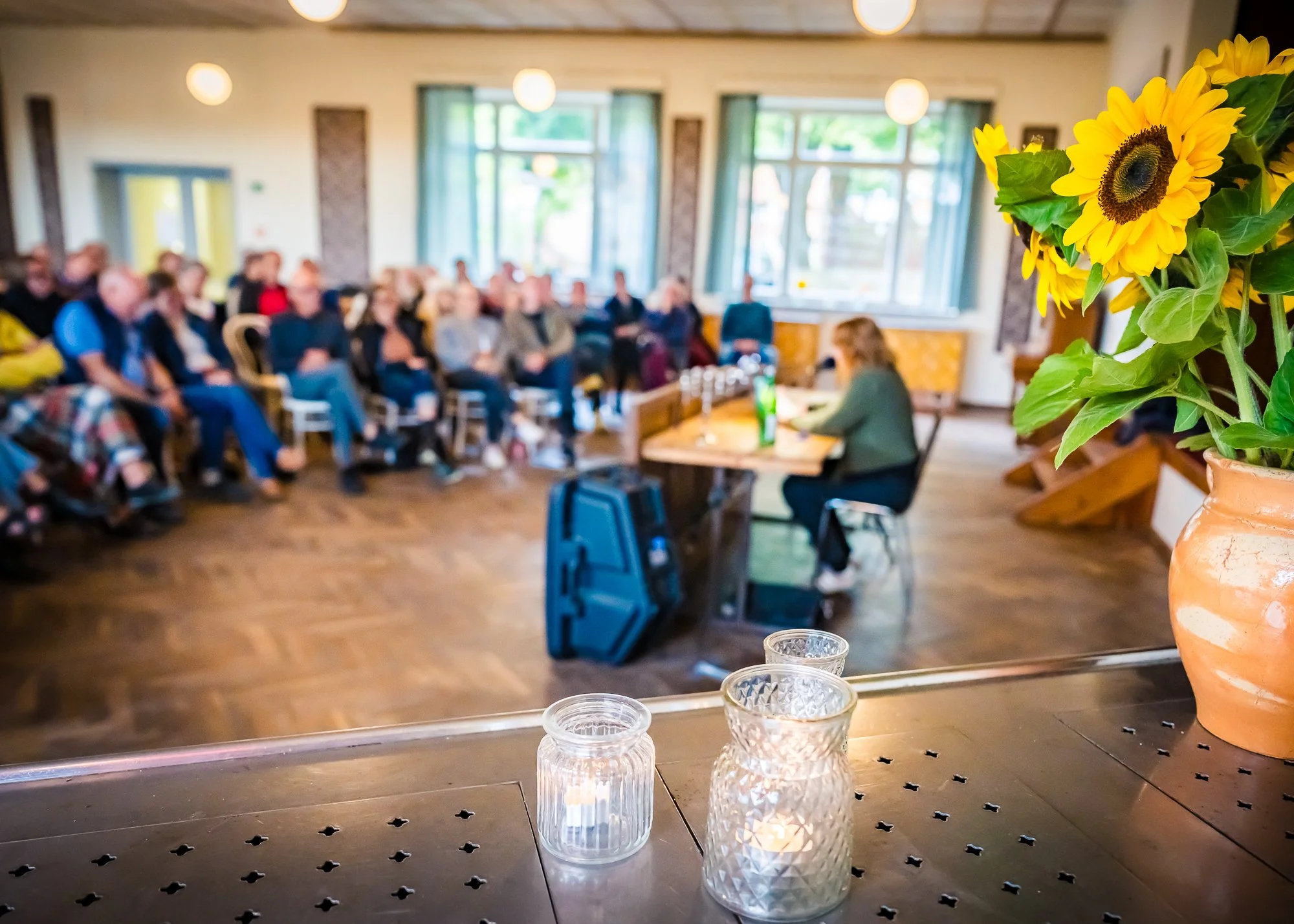 Blick auf einen Vortrag oder eine Diskussion in einem Saal, im Vordergrund sind drei Kerzen und eine Vase mit Sonnenblumen zu sehen, im Hintergrund sind mehrere Menschen, die dem Vortrag zuhören, mit Fenstern und kleinen Lampen im Raum.