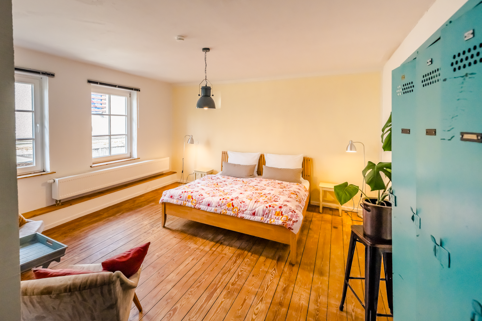 Gemütliches Schlafzimmer mit Holzfußboden, einem Doppelbett mit rosa/weißem Blumenbettbezug, zwei Nachttischen, Stehlampen, blauen Schließfächern, einer Pflanze auf einem schwarzen Hocker und zwei Fenstern.