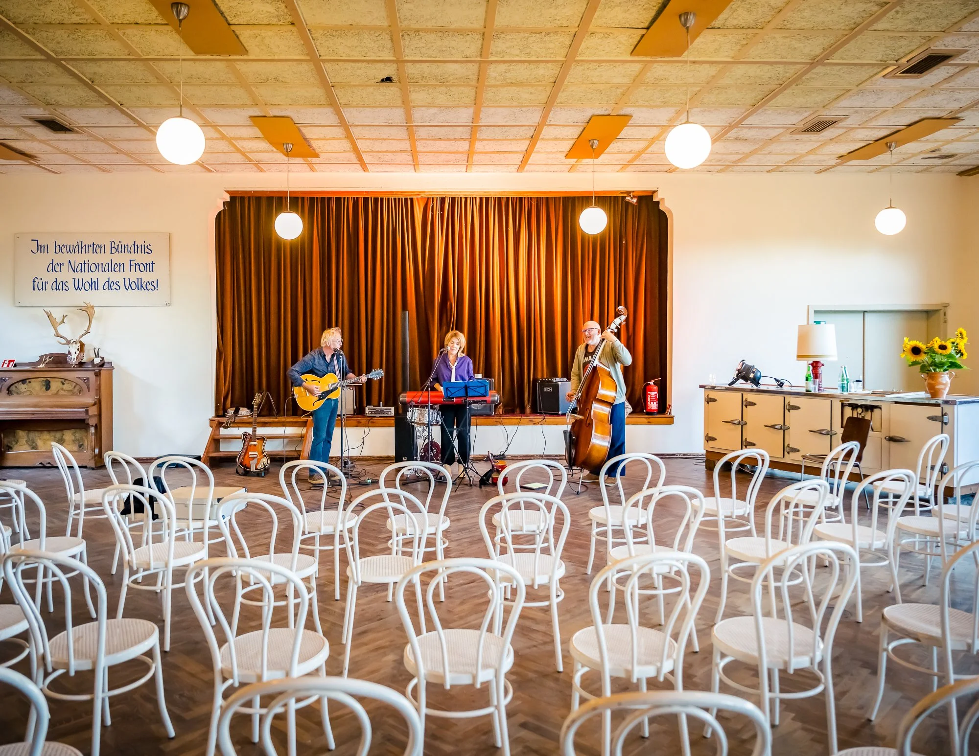 Eine Band mit drei Musikern steht auf einer kleinen Bühne in einem Saal mit weißen Stühlen. Die Musiker spielen Gitarre, Keyboard und Bass. Im Hintergrund hängt ein roter Vorhang.