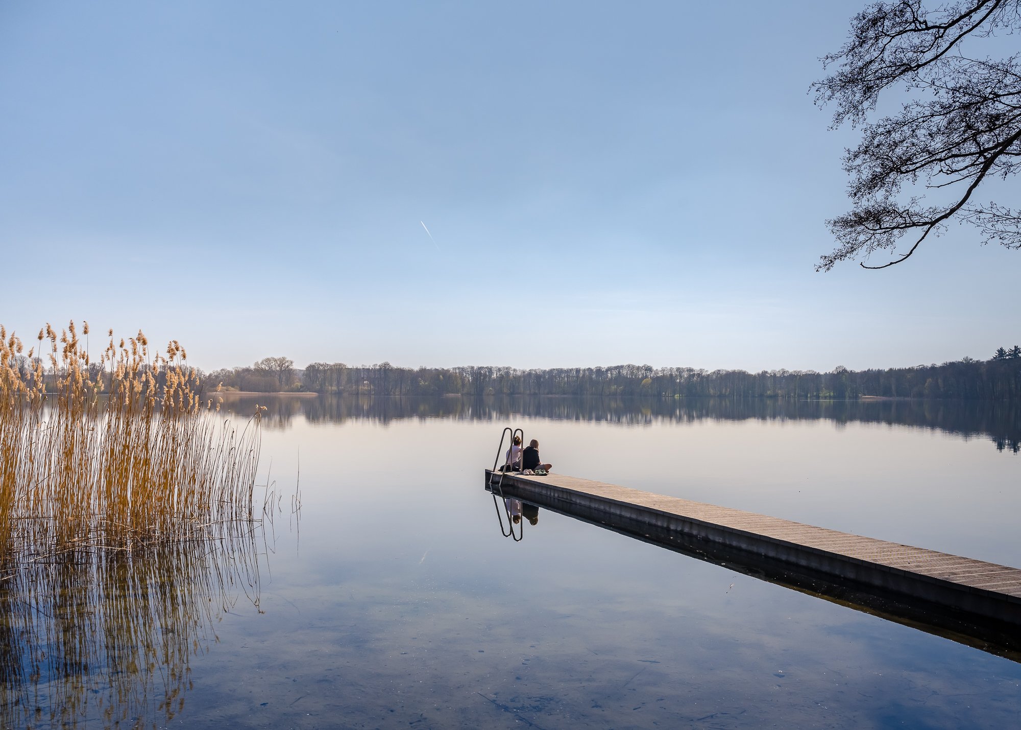 Ein ruhiger See mit einem Steg, auf dem zwei Menschen sitzen, umgeben von Bäumen und Schilf, bei klarem Himmel.