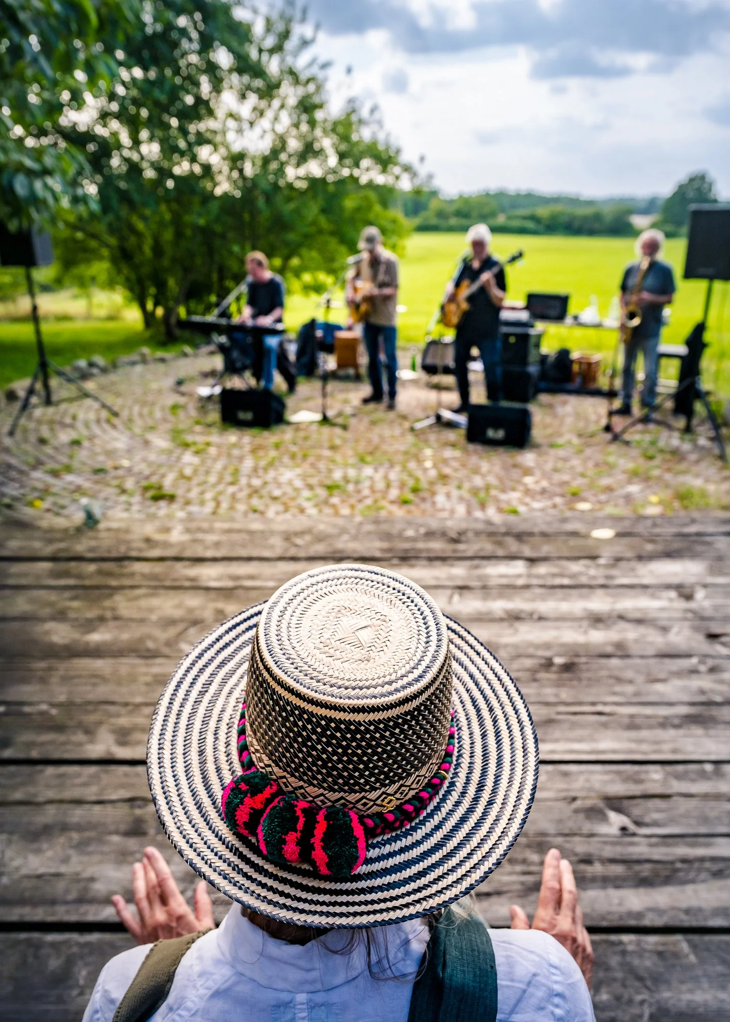 Eine Person trägt einen großen, gestreiften Hut mit bunten Bommeln und sitzt auf einer Holzplattform, während im Hintergrund eine Band live Musik macht.