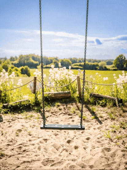 Eine leere Schaukel auf einem Sandplatz im Freien vor einer Wiesenlandschaft mit Bäumen und blühenden Sträuchern bei sonnigem Himmel.