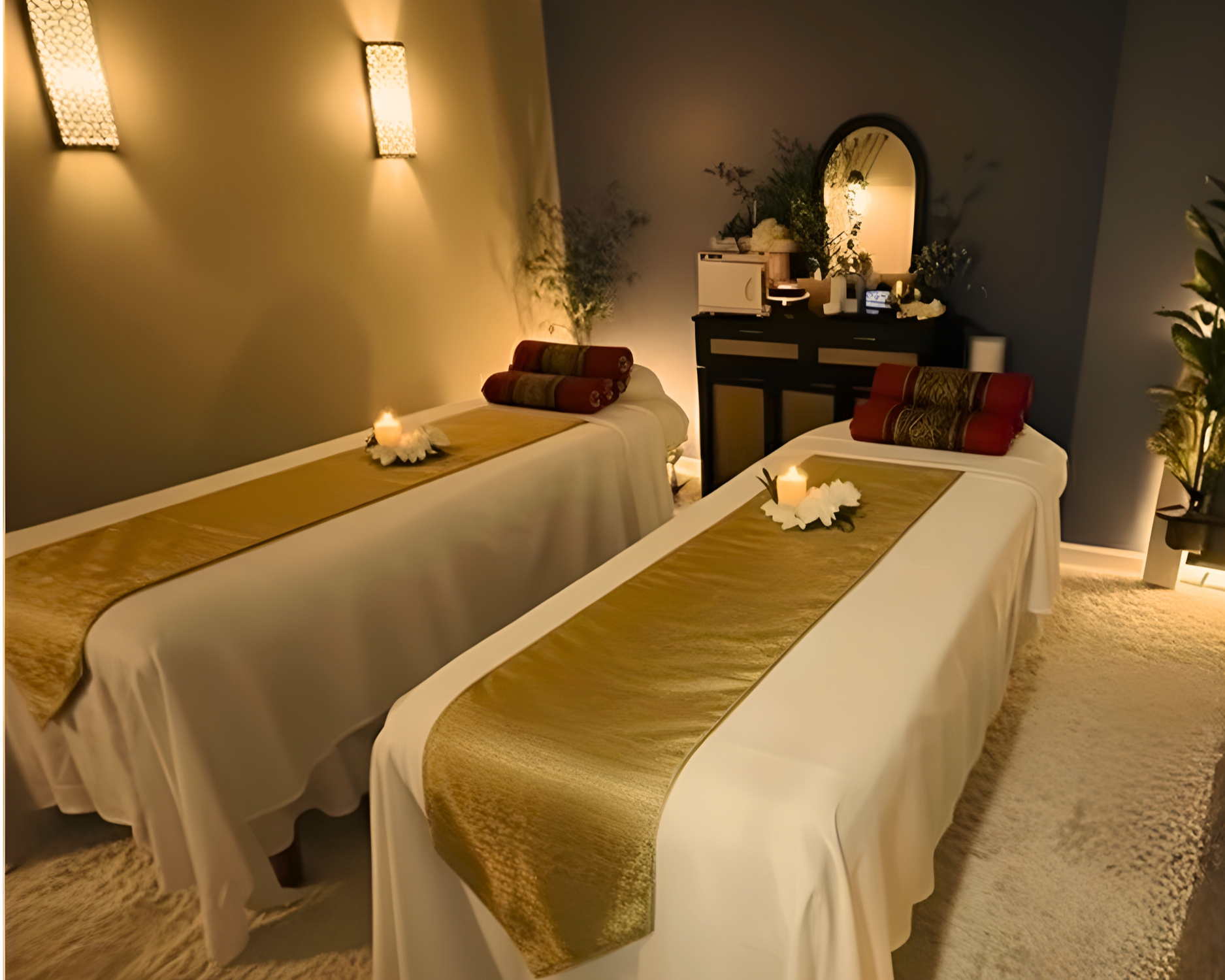 A massage therapy room with two massage tables, each draped in white sheets and gold runners, with rolled towels at the head. A great environment for massage.