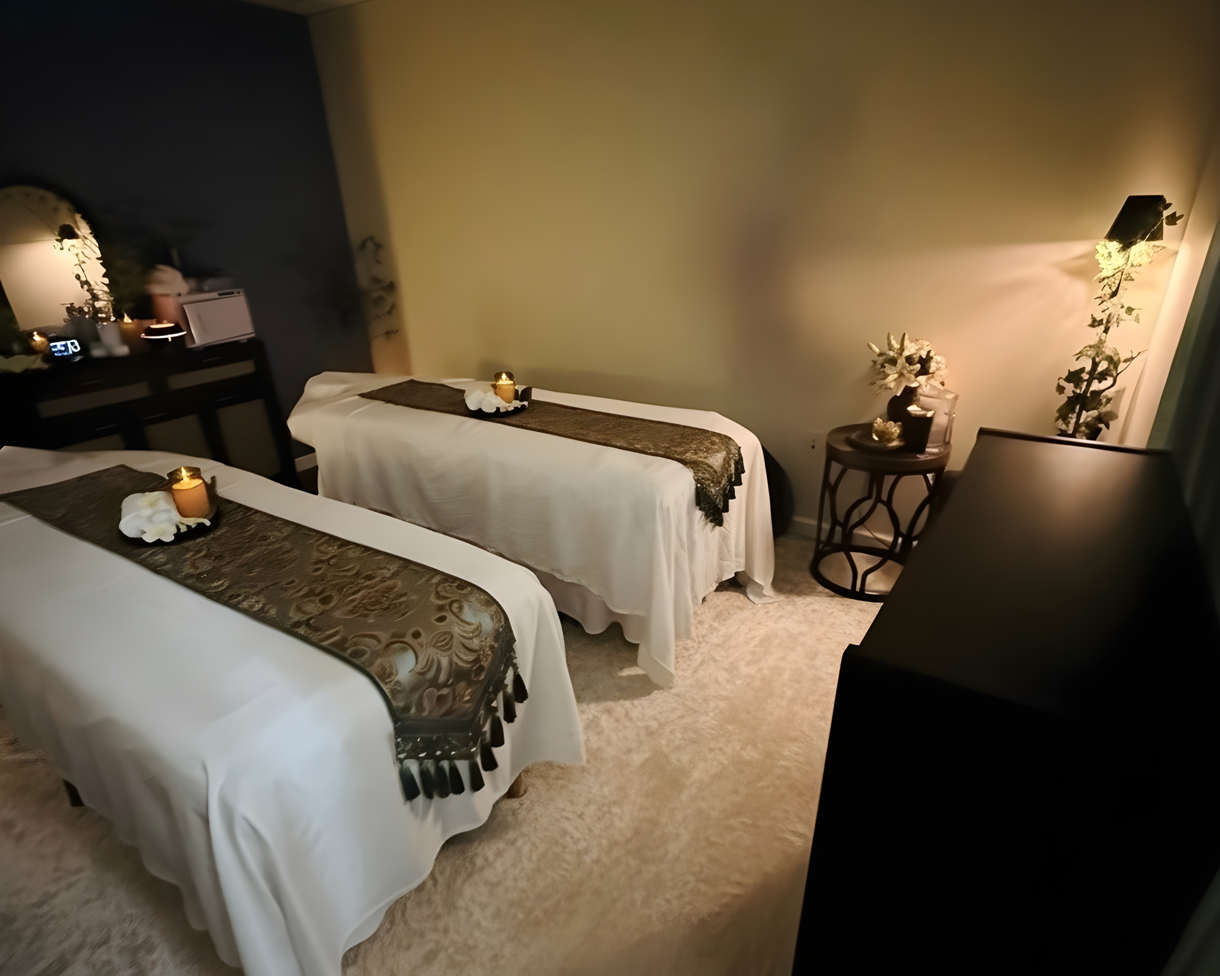 A cozy room with two massage beds covered in white sheets and decorated with brown runners, small candles, and white flowers. The room has soft lighting, a small black table with a vase of flowers, and a dark piece of furniture, with a beige carpet a