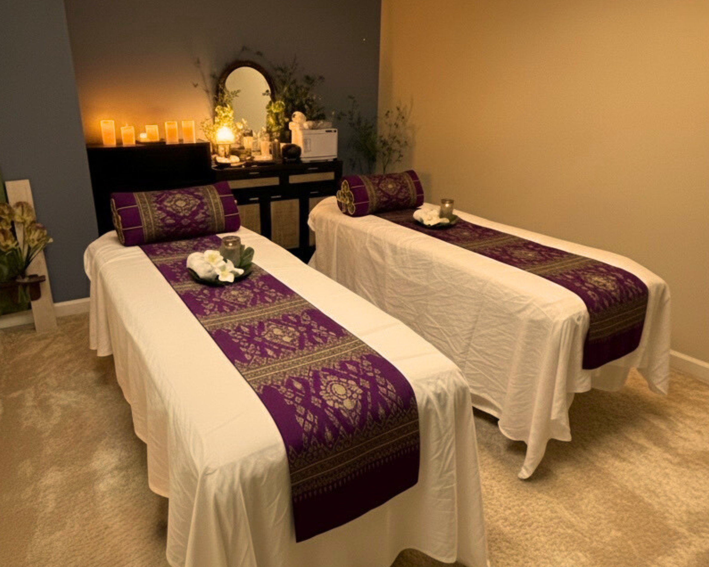 Two massage beds with purple and gold decorative runners and pillows, in a relaxing spa room with candles and plants on a black counter in the background. Experience a luxury massage.