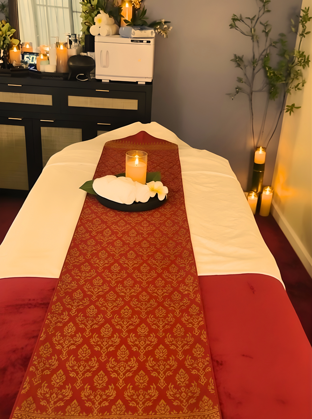 A massage or spa table with a red and gold patterned runner, lit candles, flowers, and decor, suggesting a relaxing spa or massage treatment setting.