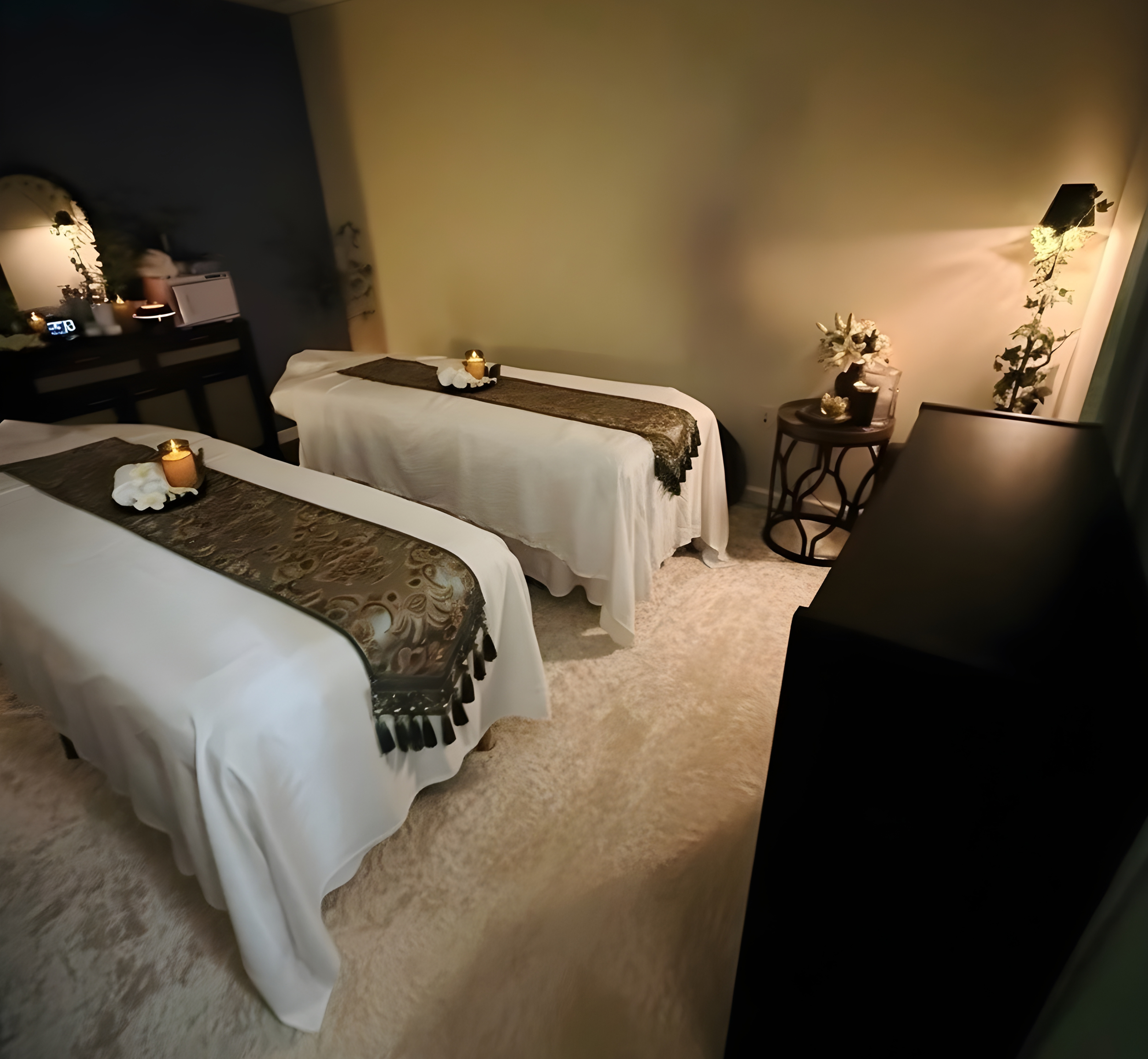 Two massage tables covered with white sheets and decorative runners, with lit candles and flower arrangements in a dimly lit, relaxing spa room.