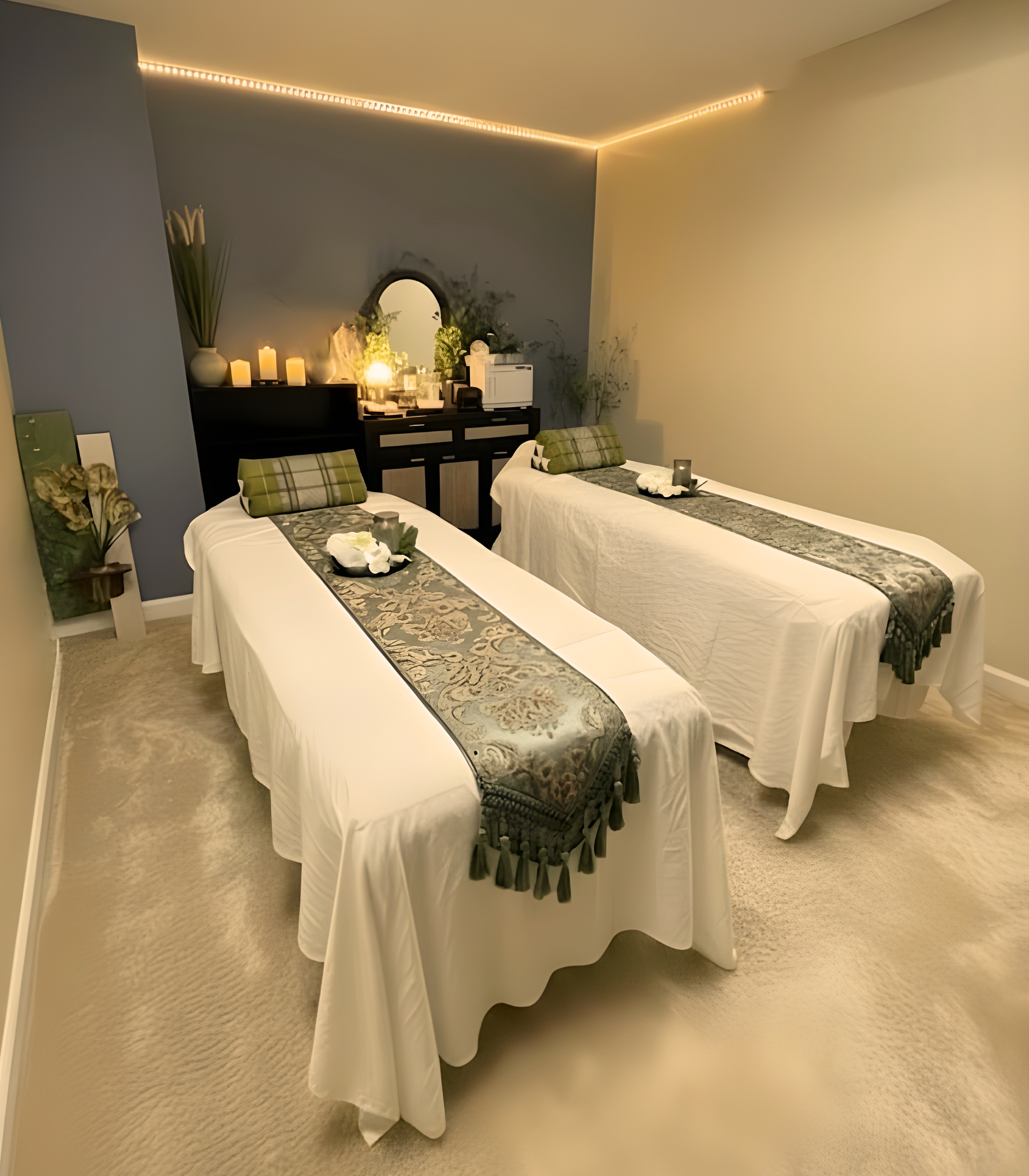 A peaceful spa room with two massage tables covered in white sheets, each adorned with a decorative runner and a plaid pillow. The room has soft lighting, candles on a black dresser, and plants for a calming atmosphere.