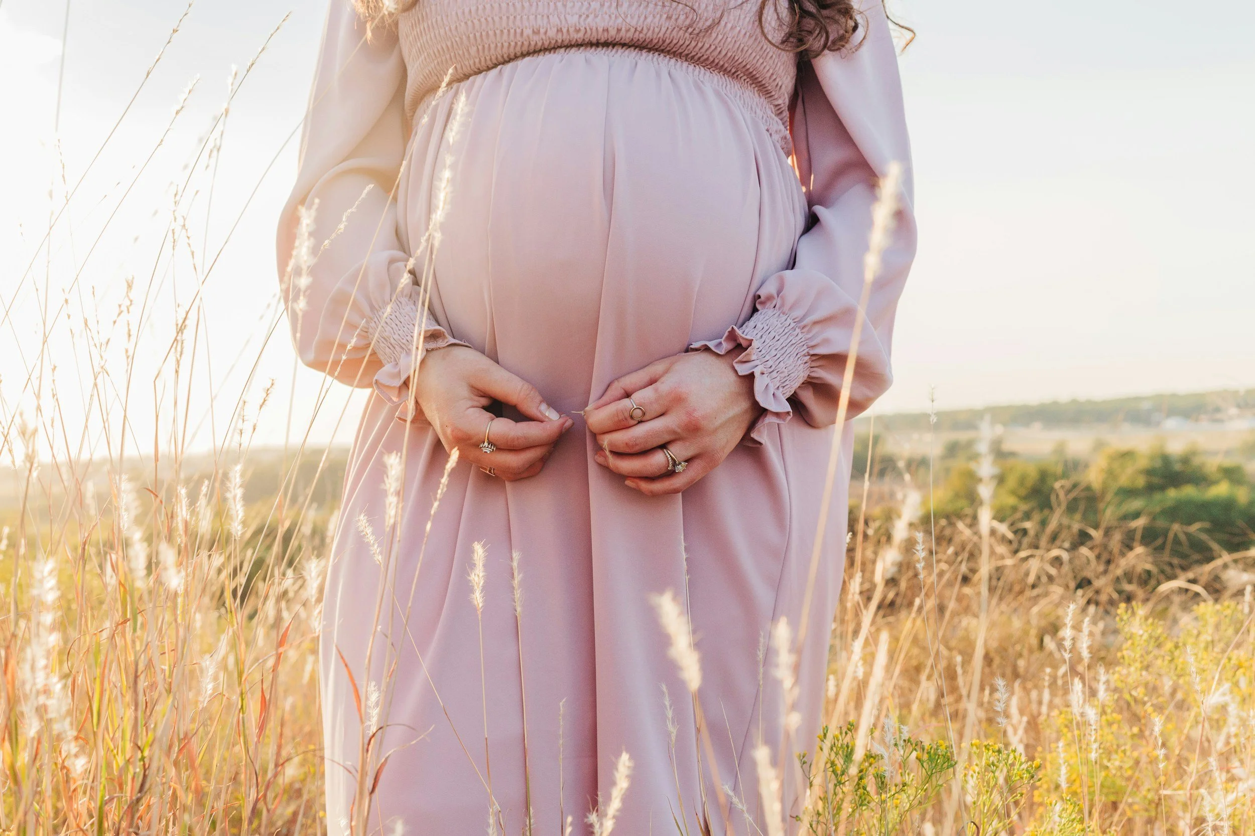 A pregnant woman wearing a pink dress and rings, standing in a field of tall grass during sunset or sunrise, with a soft and warm light.