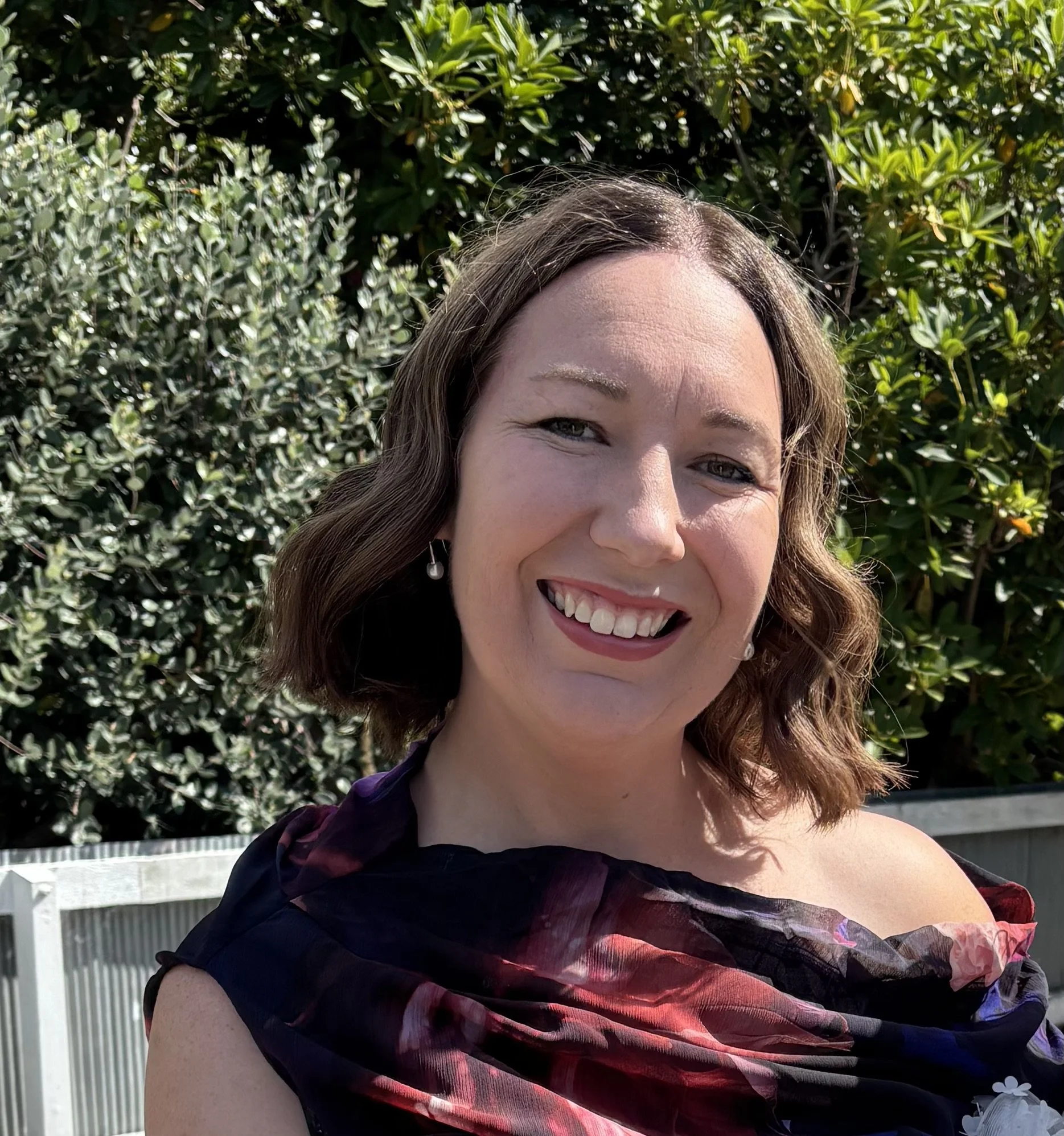 A woman smiling outdoors with green shrubbery in the background.