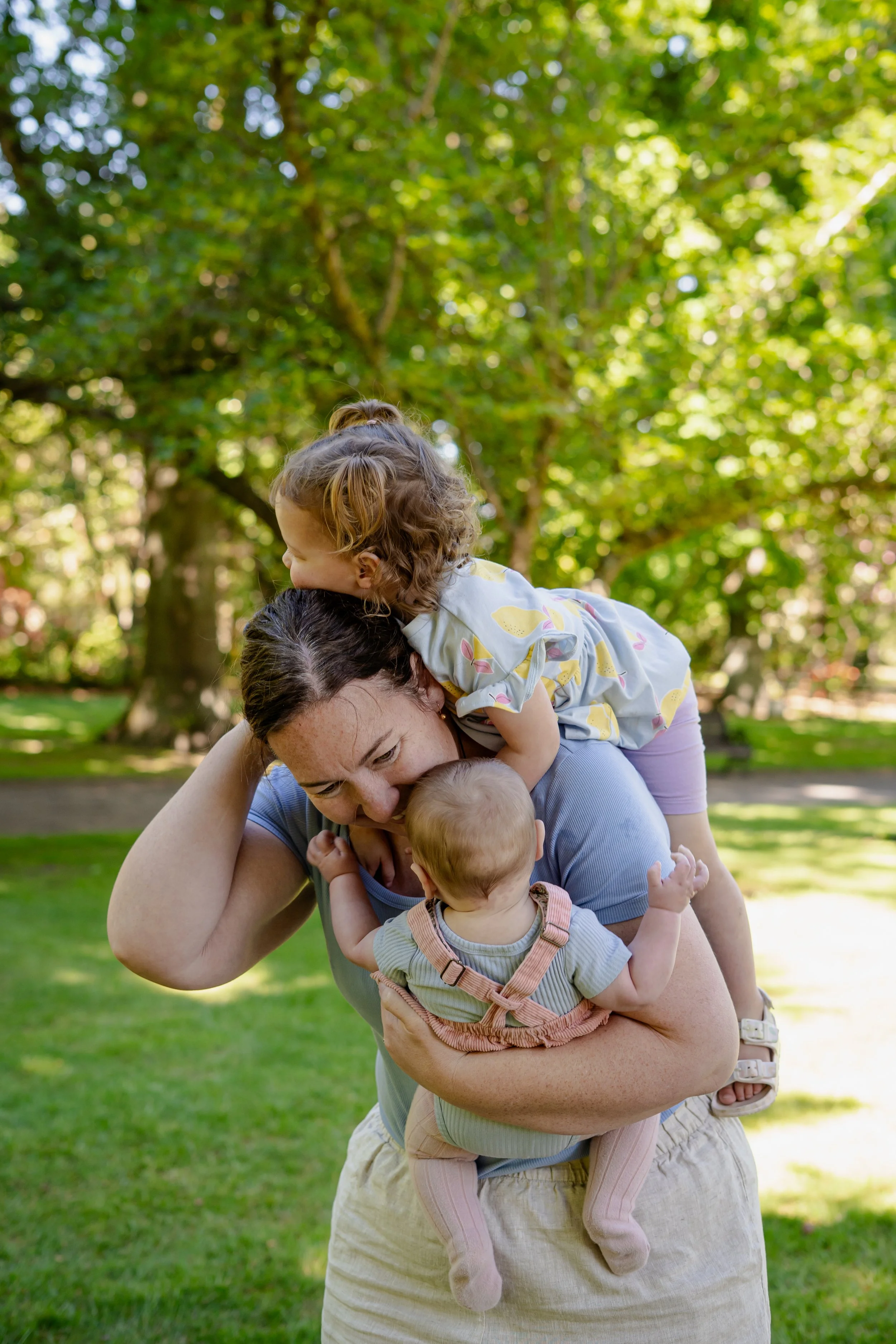 A woman holding a baby in a beige romper while a young girl with curly hair in a white dress with lemon print sits on her shoulders in a park with green trees.