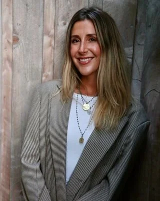 A smiling woman with shoulder-length blonde hair, wearing a plaid blazer over a white top, standing against a wooden wall.