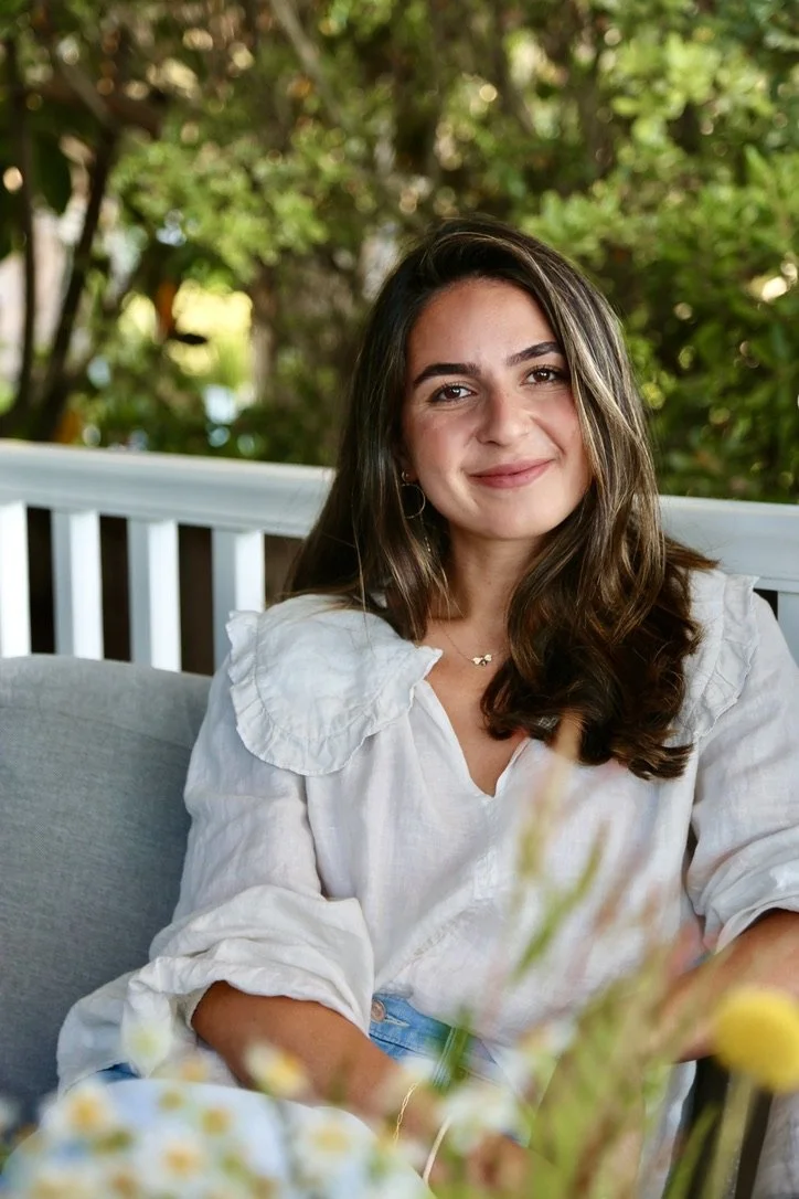 A young woman with long brown hair and a white blouse sitting outdoors on a white bench, smiling at the camera, with greenery in the background.
