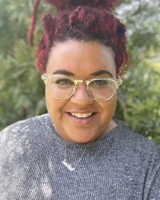 A woman with red curly hair, wearing glasses, a gray sweater, and a necklace, smiling outdoors with greenery in the background.
