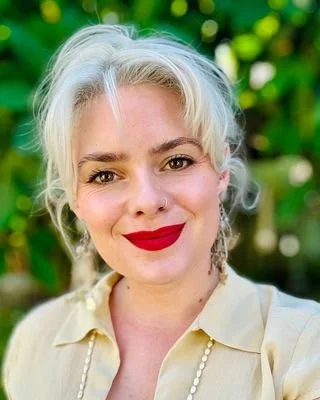 A woman with platinum blonde hair, red lipstick, and a nose piercing, smiling outdoors with a blurred green background.