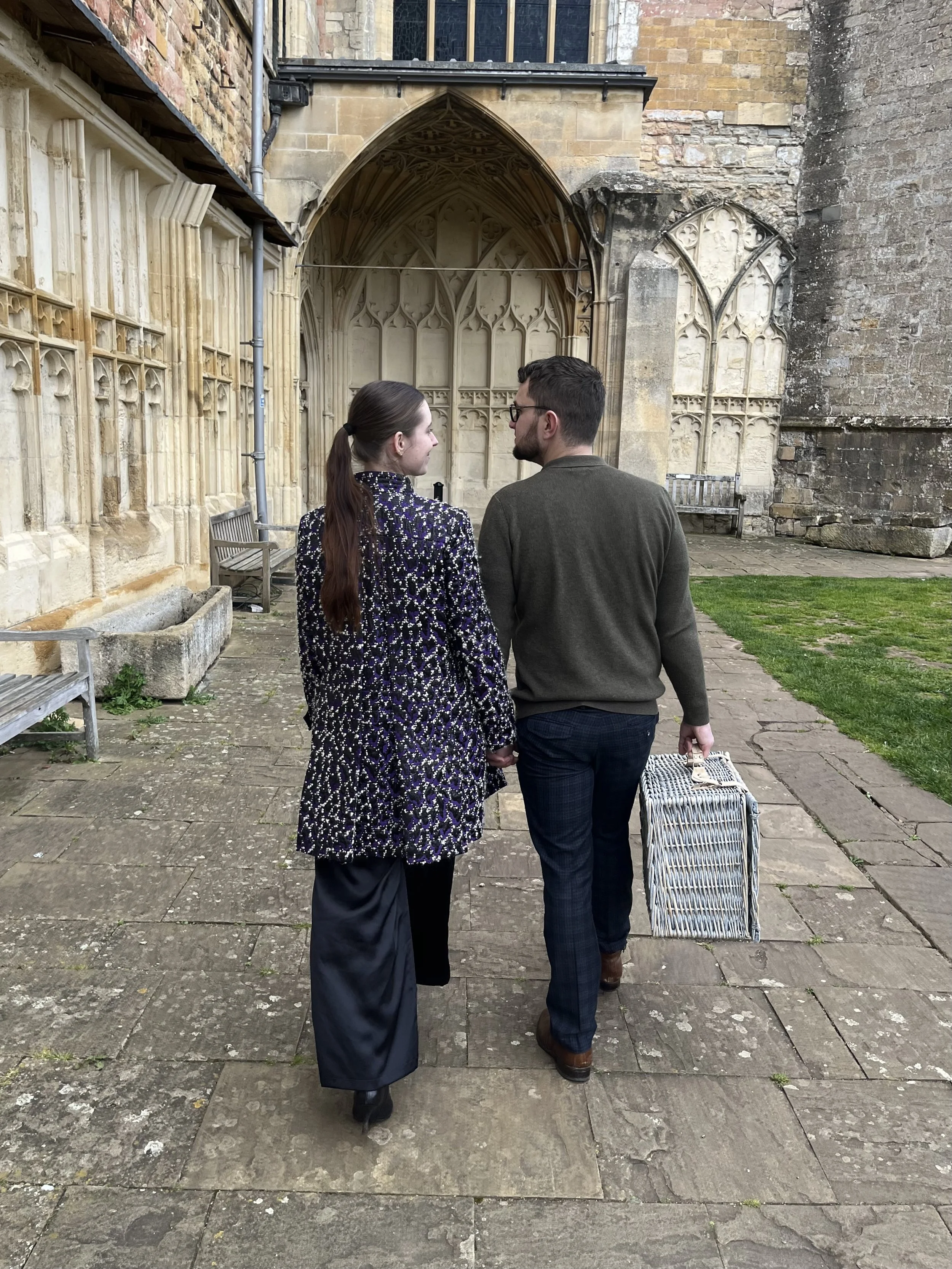 A young couple walking hand in hand outside of a historic building with Gothic architecture. The woman has long dark hair tied in a ponytail and is wearing a patterned coat, while the man has short dark hair, glasses, a brown sweater, and plaid pants, carrying a striped suitcase.