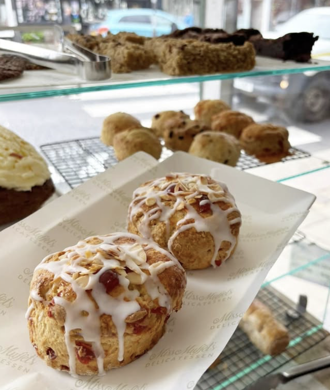 Close-up of two scones with icing and nuts on a white tray inside a bakery. Behind them, a glass display case shows various pastries, cookies, and baked goods, with some racks of cookies and brown bread on top. Outside the window, cars are visible.