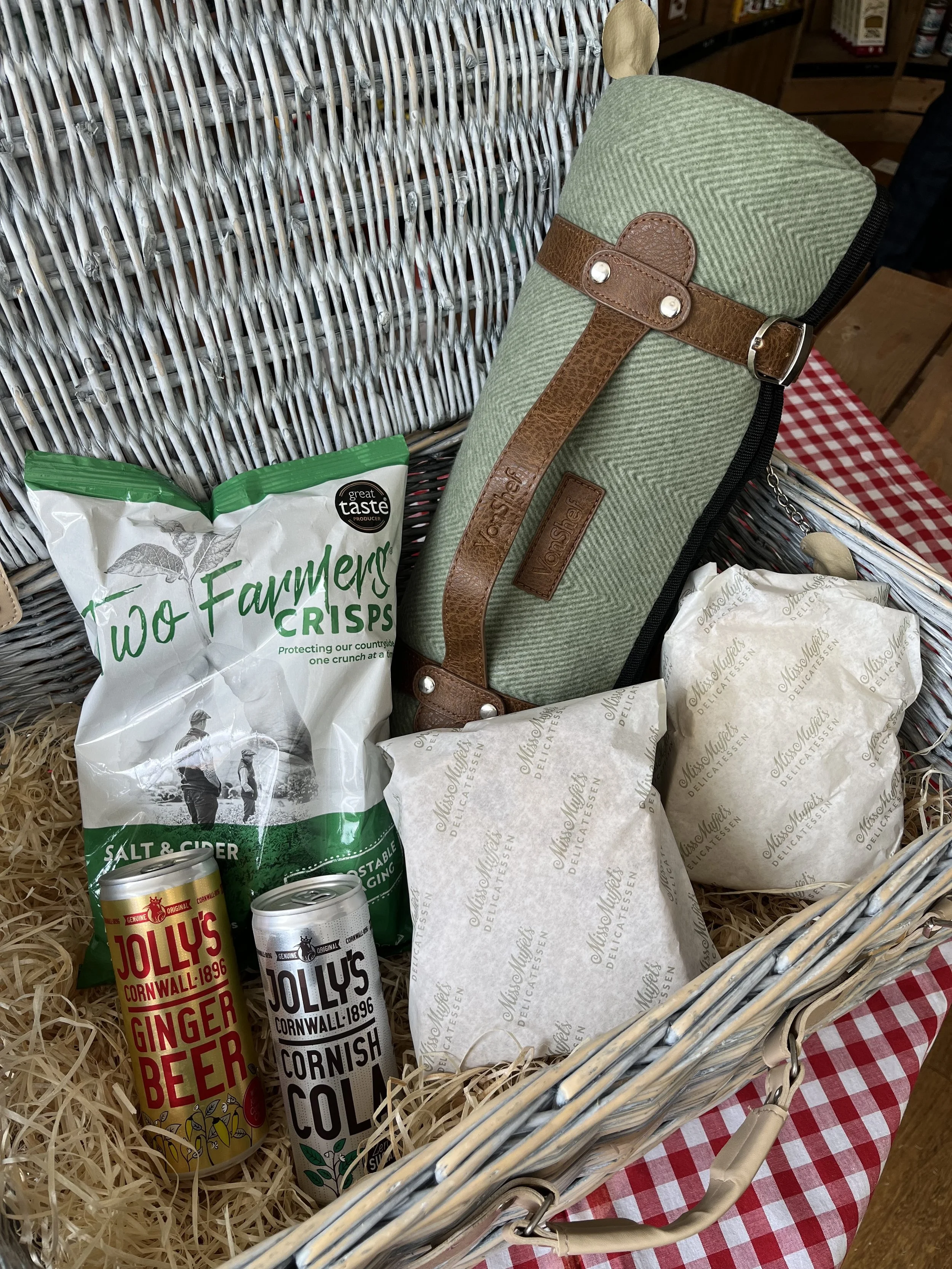 A gift basket containing two cans of Jolly's Cornwall ginger beer and cornish cola, a green and brown toiletry bag with leather strap, and two wrapped sandwiches inside a wicker basket with shredded paper, placed on a red and white checkered tablecloth.
