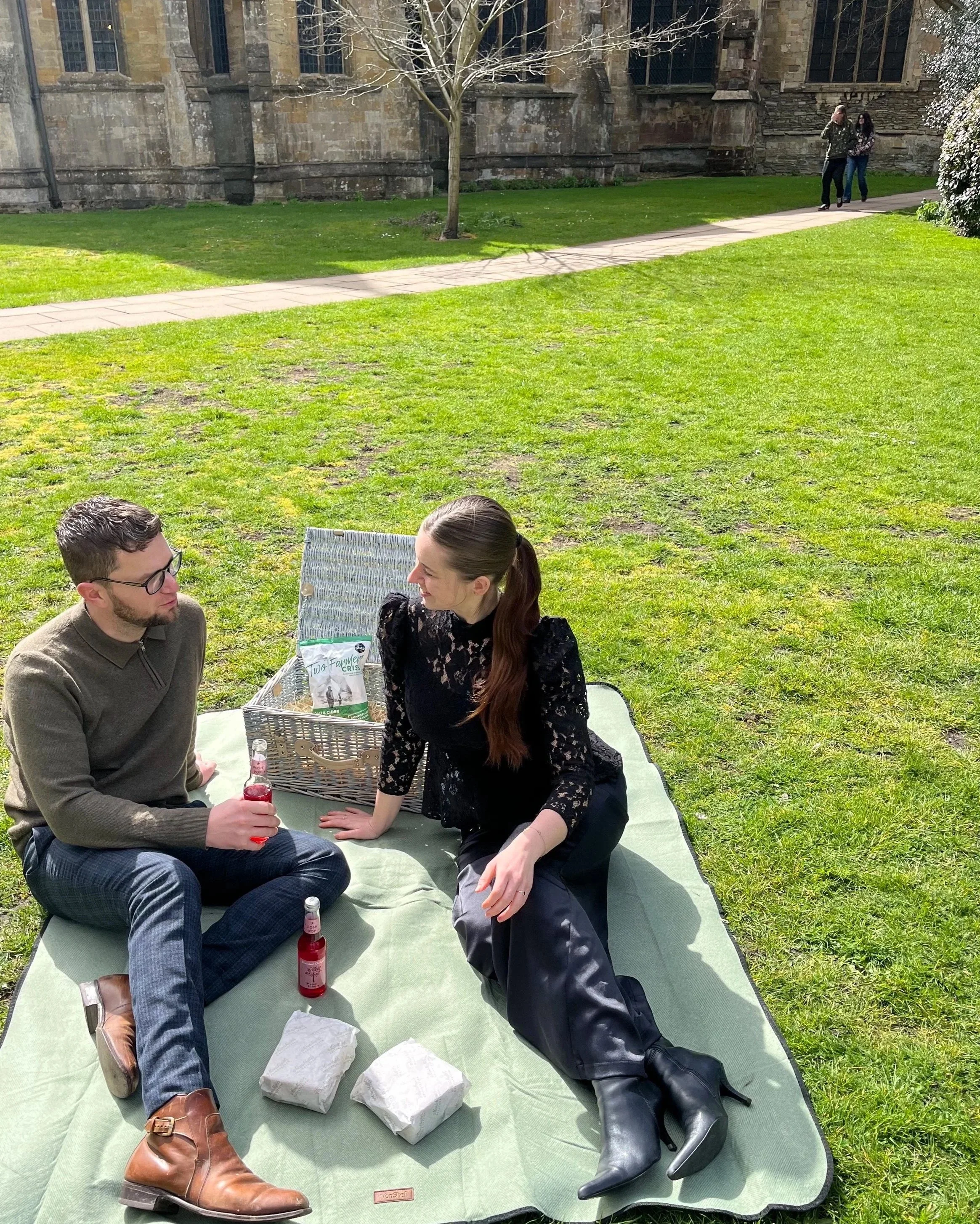 A man and woman having a picnic at Tewkesbury Abbey. They are sitting cross-legged and talking, with drinks in their hands. There are sandwiches wrapped in paper on the mat and a picnic basket beside them.