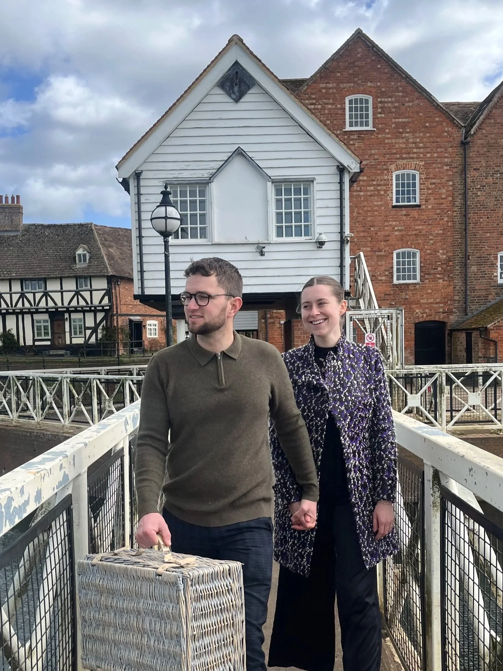 A man and woman walking hand-in-hand on a bridge, holding a picnic basket, near Tewkesbury canal.
