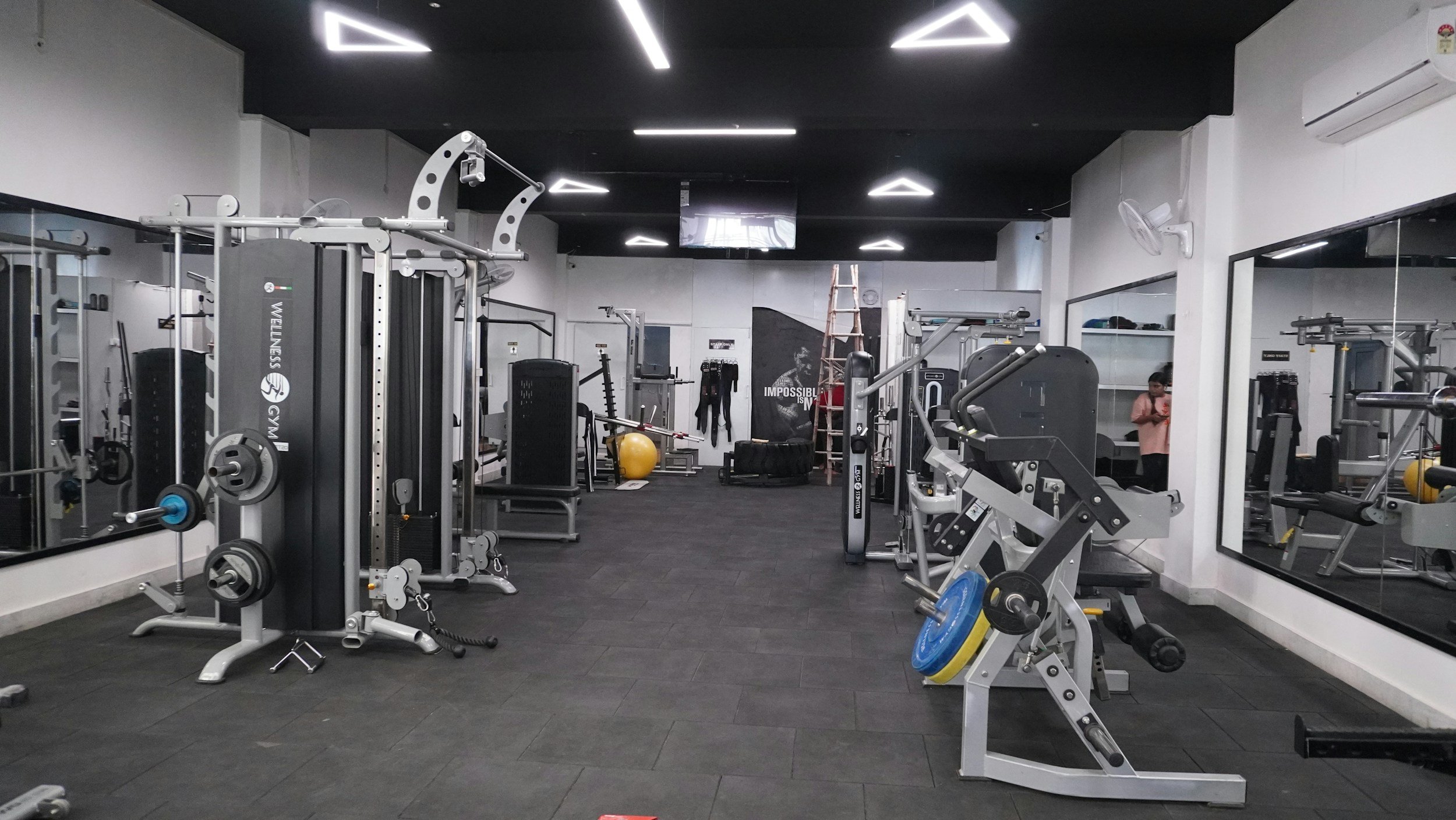 Empty fitness gym with workout equipment, weight machines, free weights, yellow exercise ball, and a person in pink shirt using a phone.