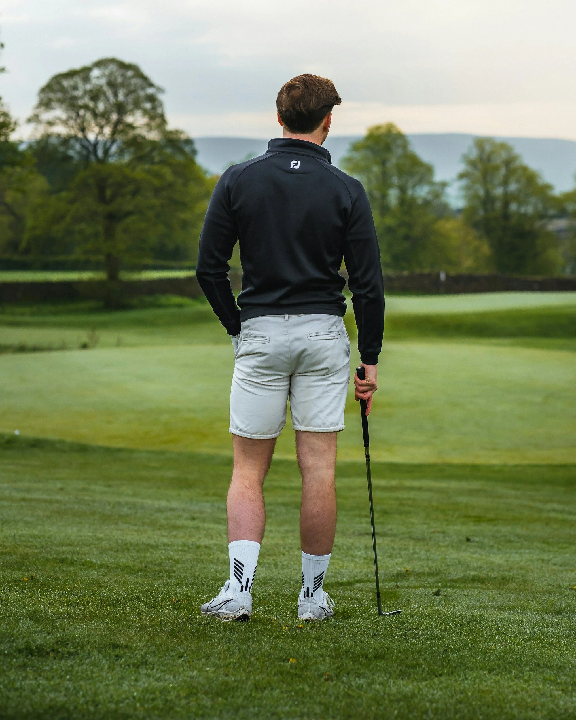 A man standing on a golf course, holding a golf club, dressed in a black jacket, white shorts, and white golf shoes, with trees and hills in the background.
