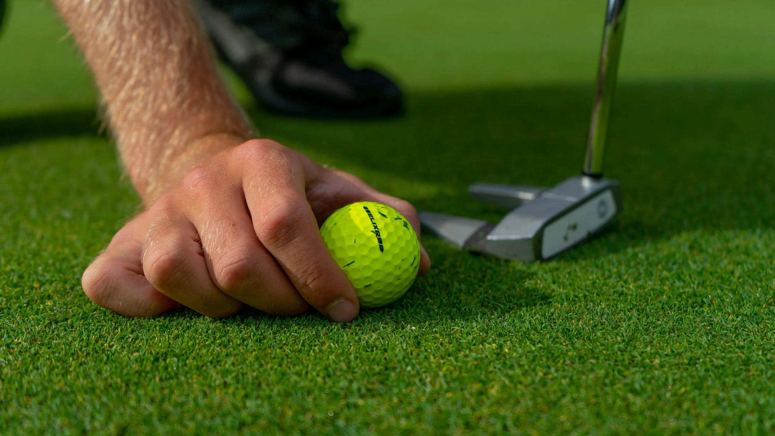 Close-up of a person lining up a yellow golf ball on a putting green with a golf club nearby.