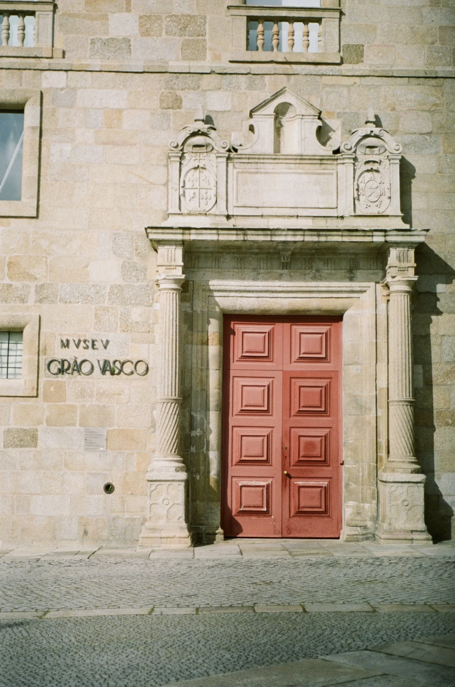 Porta vermelha de madeira em uma fachada de pedra, com detalhes arquitetônicos clássicos e uma placa que diz "Museu Grão Vasco"