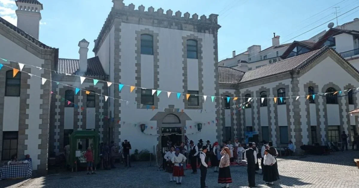 Pessoas vestidas com roupas tradicionais em uma praça em frente a um prédio antigo decorado com bandeirinhas coloridas, em uma celebração ao ar livre.