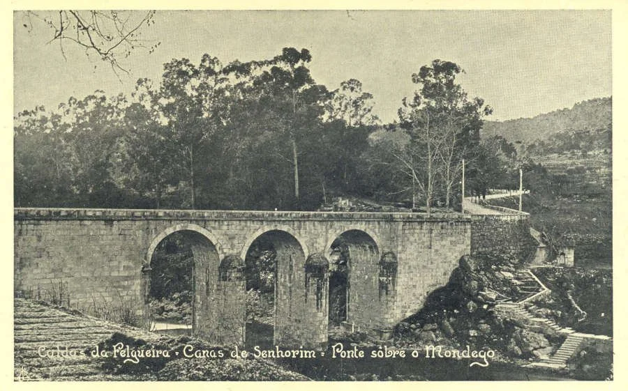 Imagem antiga de um ponte de pedra com arcos sobre um rio ou vale, cercada por árvores e vegetação, com uma paisagem de colinas ao fundo.