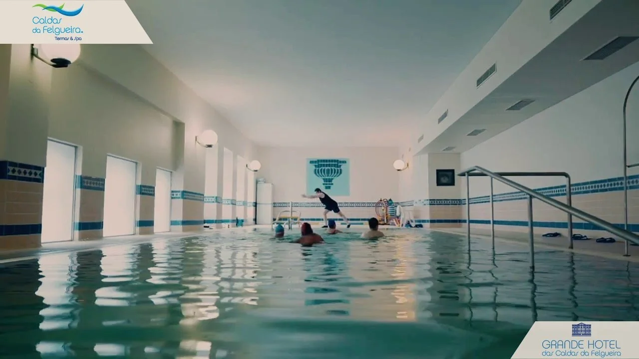 Piscina interna em um hotel, com uma pessoa ensinando ou ajudando crianças na água. paredes brancas com janelas altas e iluminação suave, decoração minimalista com alguns quadros na parede. Logo no canto superior esquerdo, texto em português e azul "