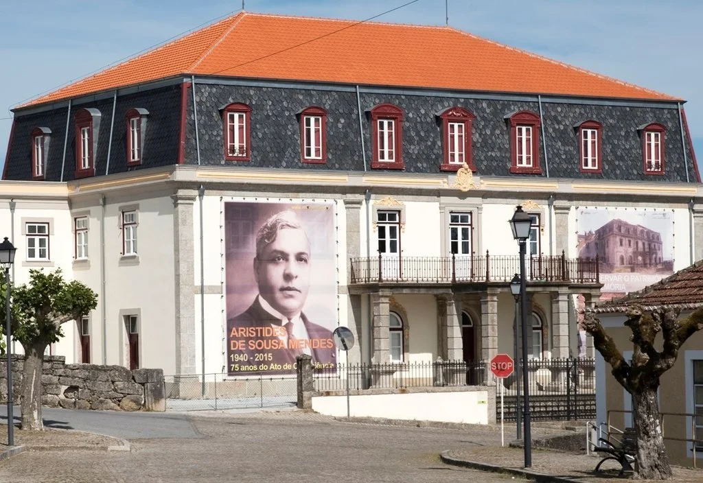 Prédio antigo com telhado de telhas vermelhas, janelas altas e uma grande imagem de Aristedes de Sousa Mendes na fachada, em um evento que comemora seus 75 anos desde a assinatura do Ato de Varginha.