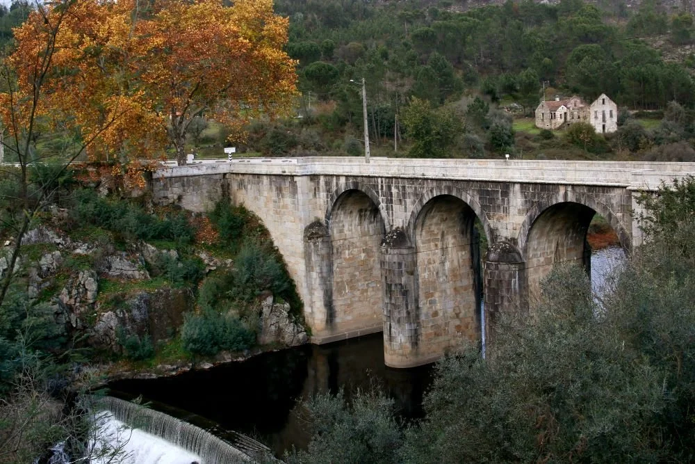 Ponte de pedra antiga de três arcos sobre um rio, com árvores ao redor e casas ao fundo em uma paisagem rural.