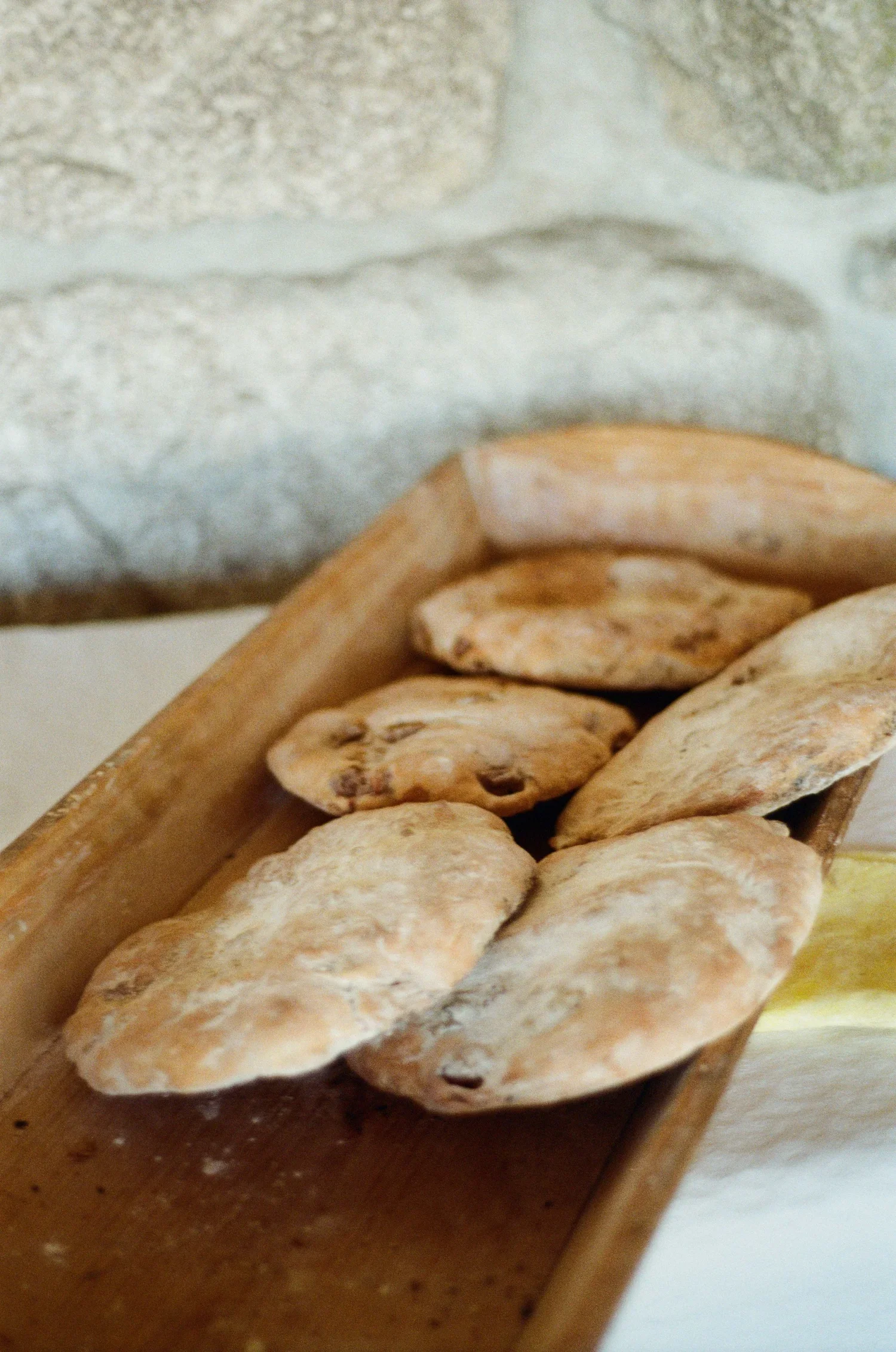 Pedaços de pão sírio ou focaccia em uma bandeja de madeira, com uma parede de pedra ao fundo.