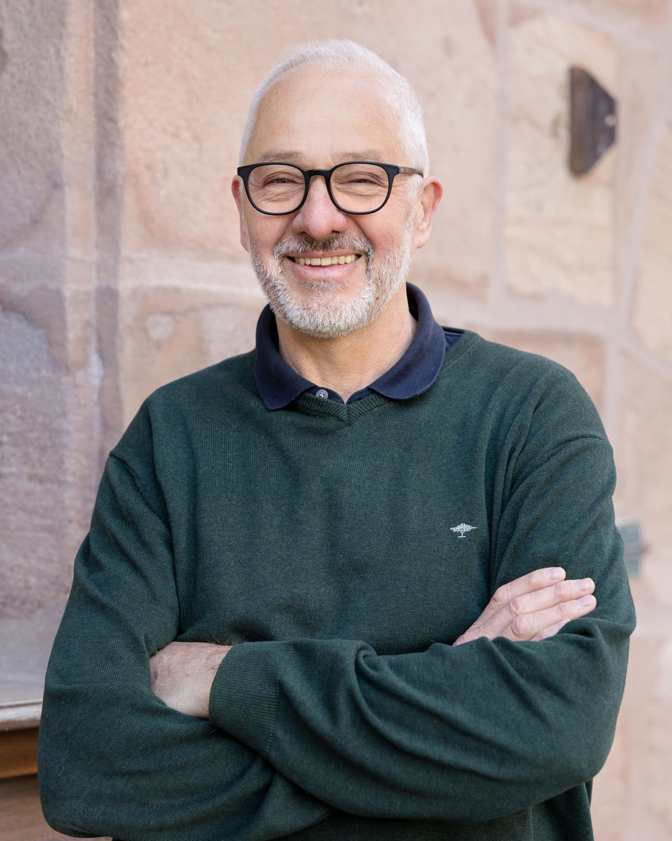 Ein Mann mit Brille, grauem Haar und Bart, lächelnd, trägt einen grünen Pullover mit Kragen, steht vor einem Steinmauer.