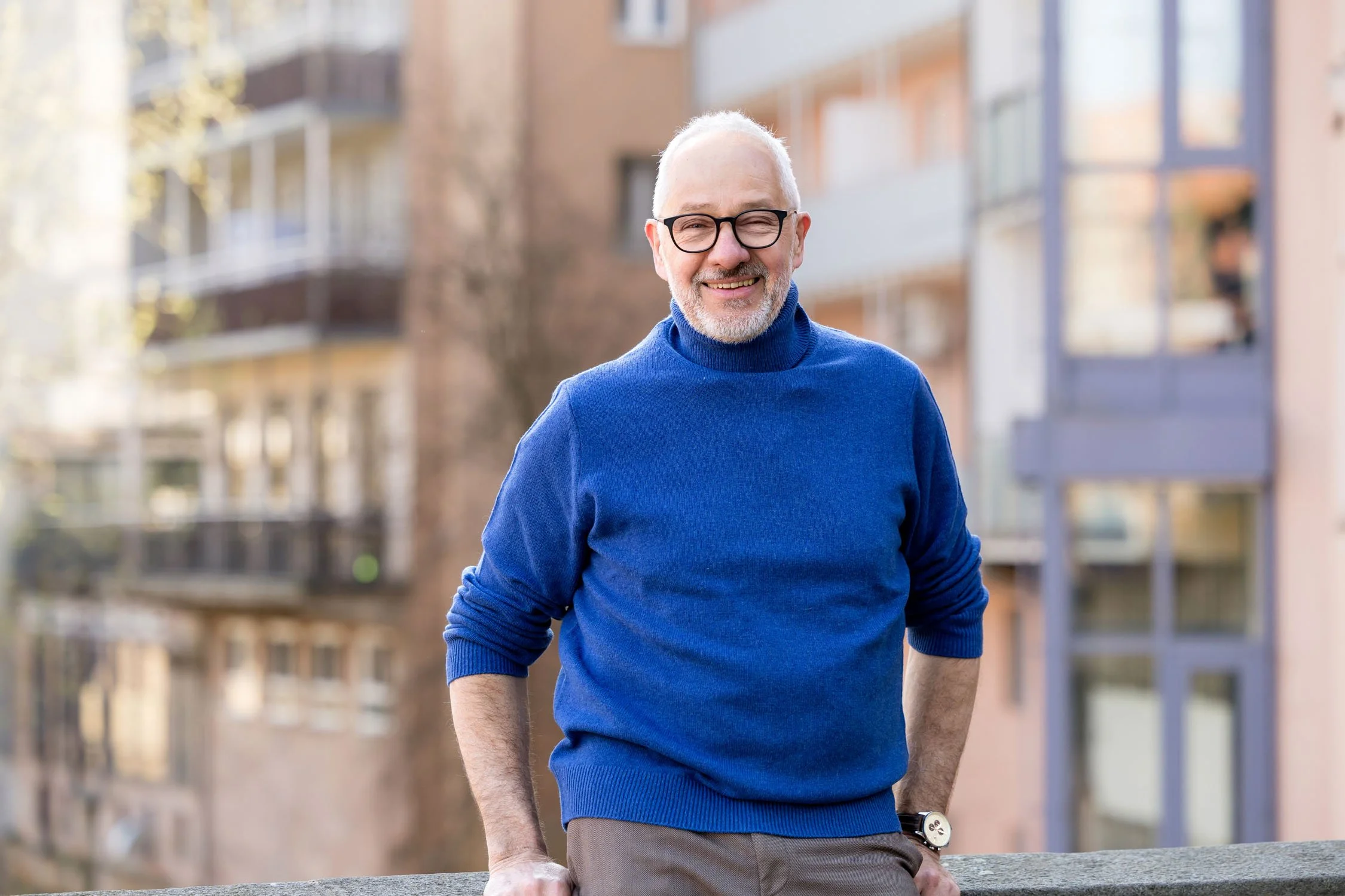 Ein Mann mit Brille, grauem Bart und blonden Haaren, der draußen vor einem mehrstöckigen Gebäude steht. Er trägt einen blauen Pullover und lächelt in die Kamera.