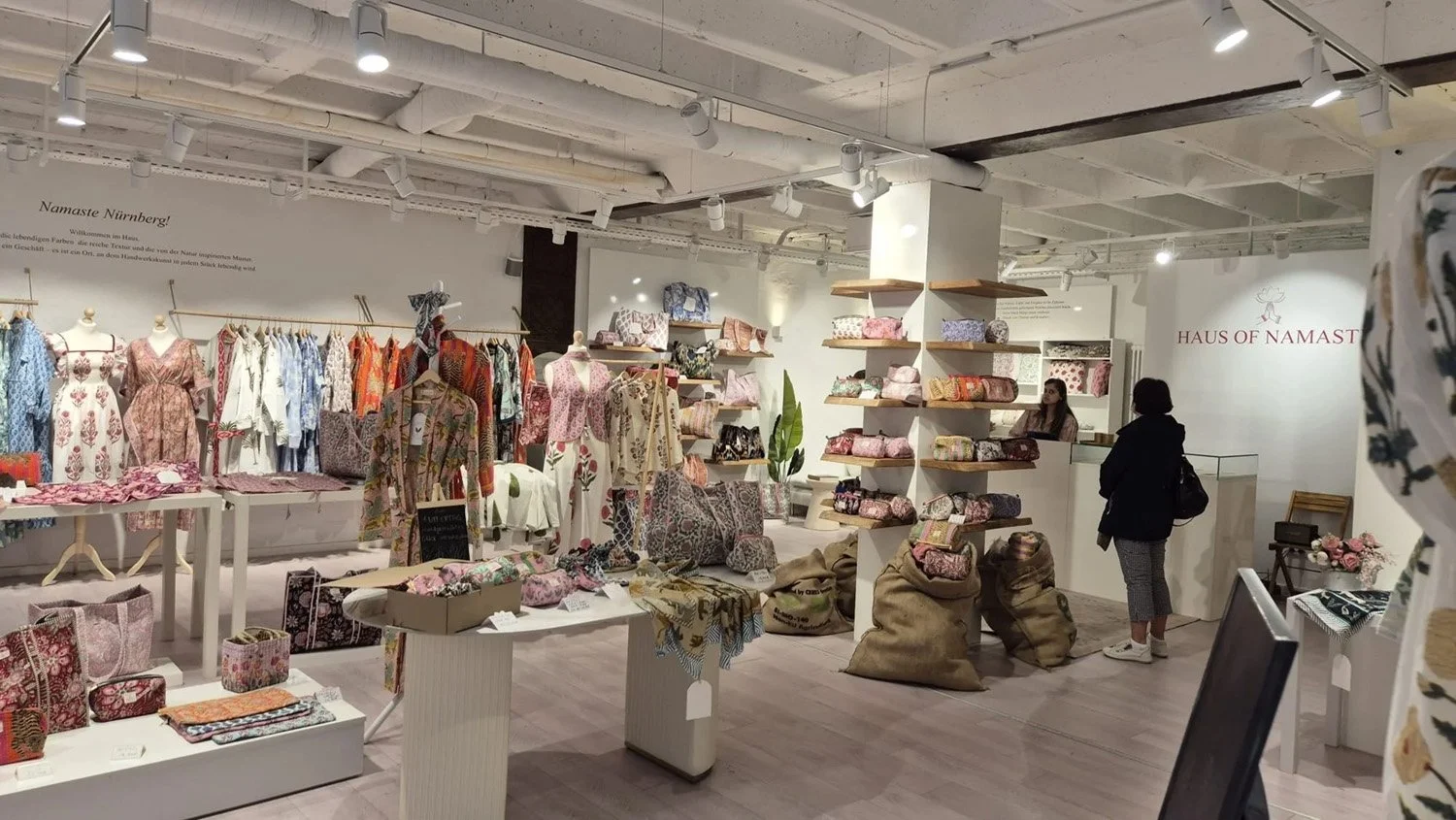 Ein Geschäft mit mediterran inspirierten Textilien und Accessoires, das Kleidung, Kissen, Taschen und Stoffe auf weißen Regalen und Tischen präsentiert. Zwei Kunden betrachten die Produkte, im Hintergrund eine Mitarbeiterin an der Kasse, Wände mit Beschriftungen 'Namaste Nürnberg!' und 'Haus of Namaste'.