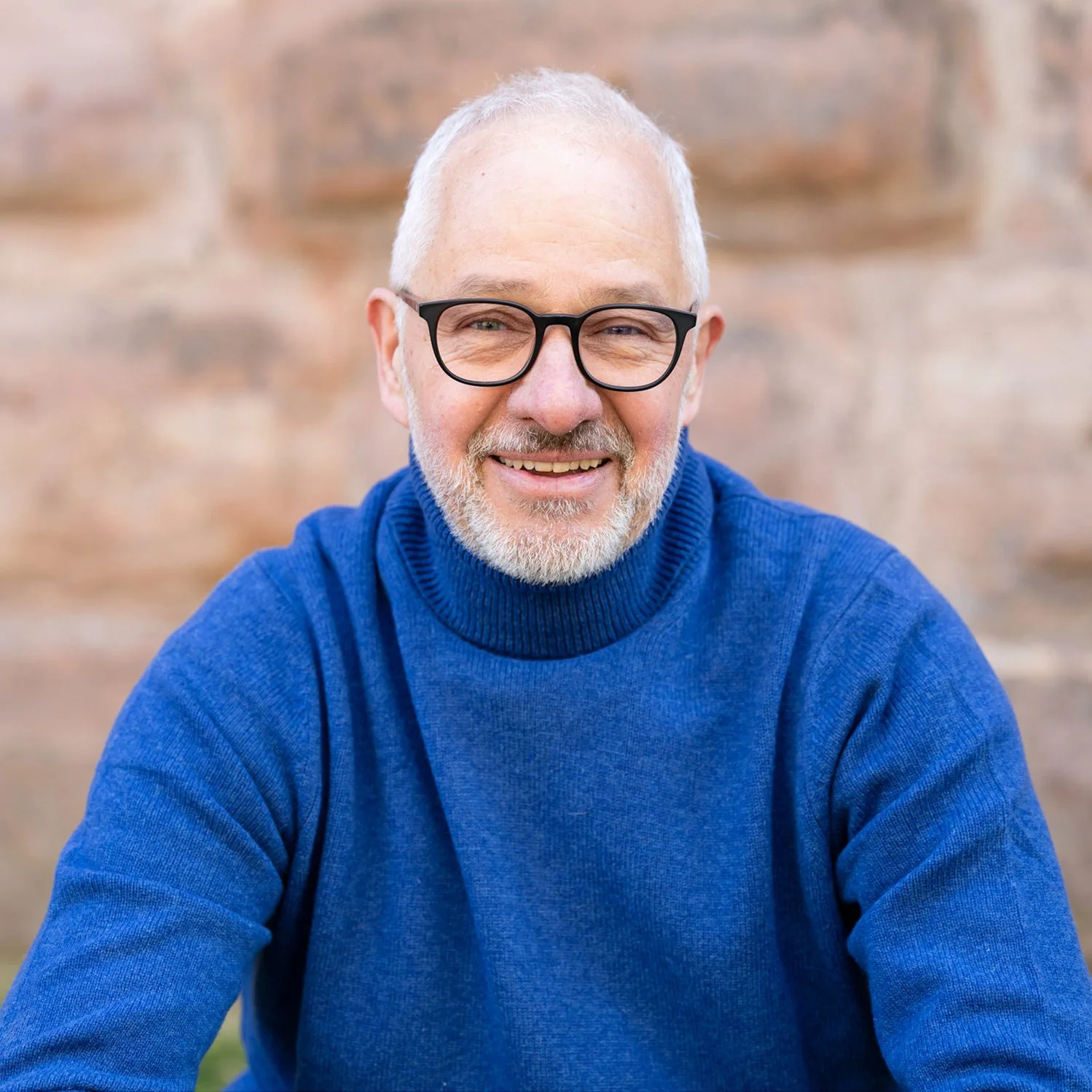 Mann mit Brille und weißem Bart, trägt einen blauen Rollkragenpullover, sitzt vor einem Backsteinwand, lächelt in die Kamera.