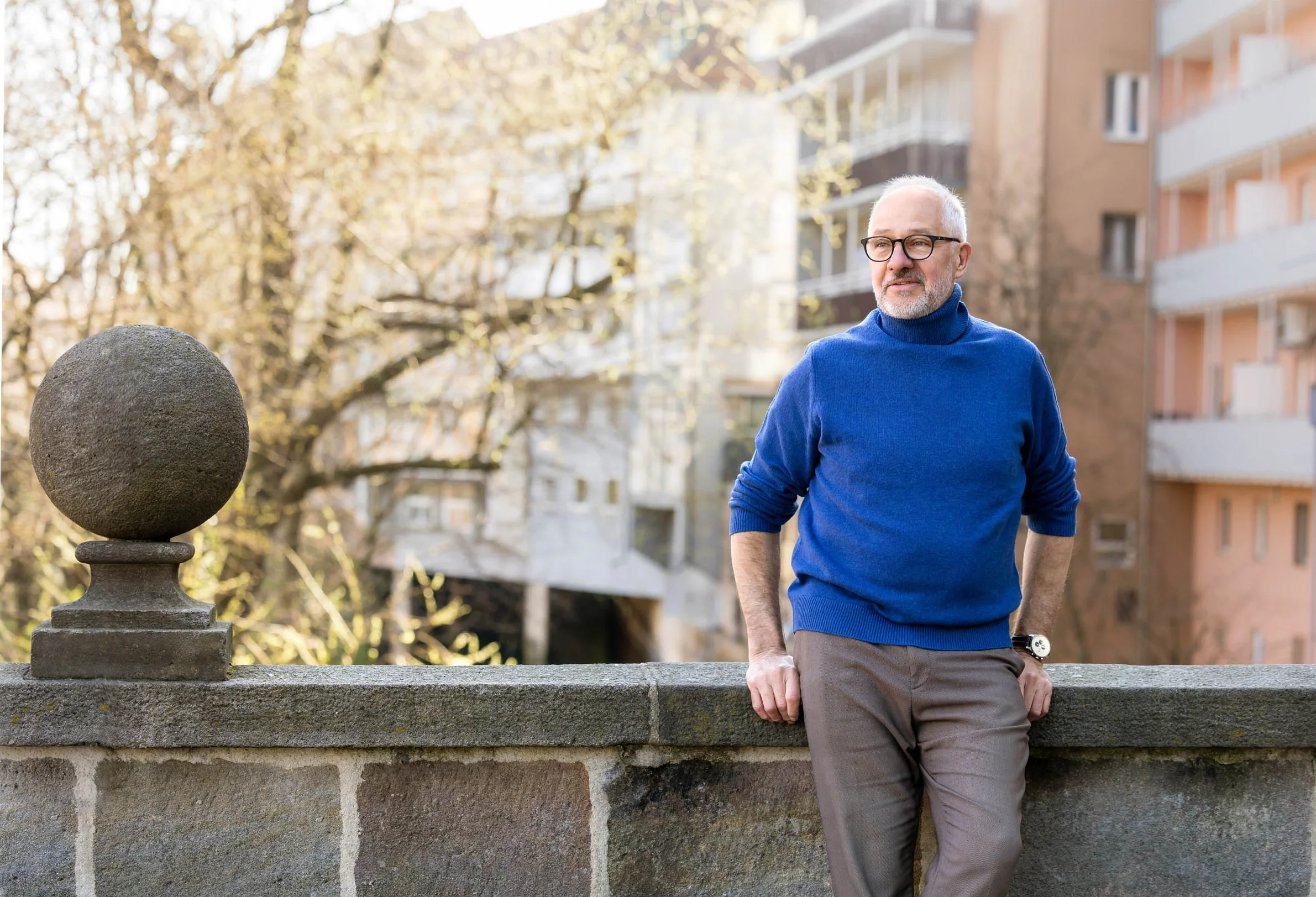 Ein Mann mit Brille, grauem Bart und hellgrauem Haar, der einen blauen Rollkragenpullover trägt, lehnt an einer Steinmauer, im Hintergrund Bäume und mehrstöckige Wohnhäuser, sonniger Tag.