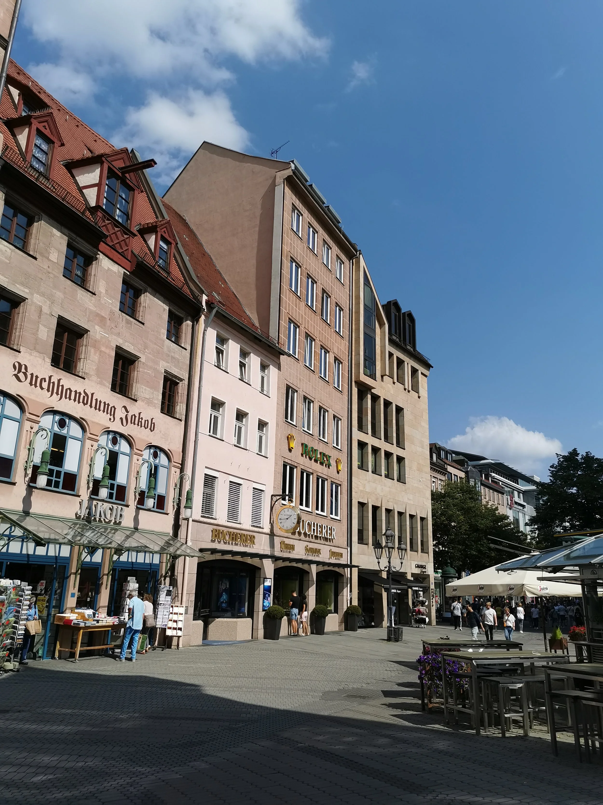 Stadthaus in einer belebten Fußgängerzone mit Geschäften, Café-Terrasse, Wolken am blauen Himmel.