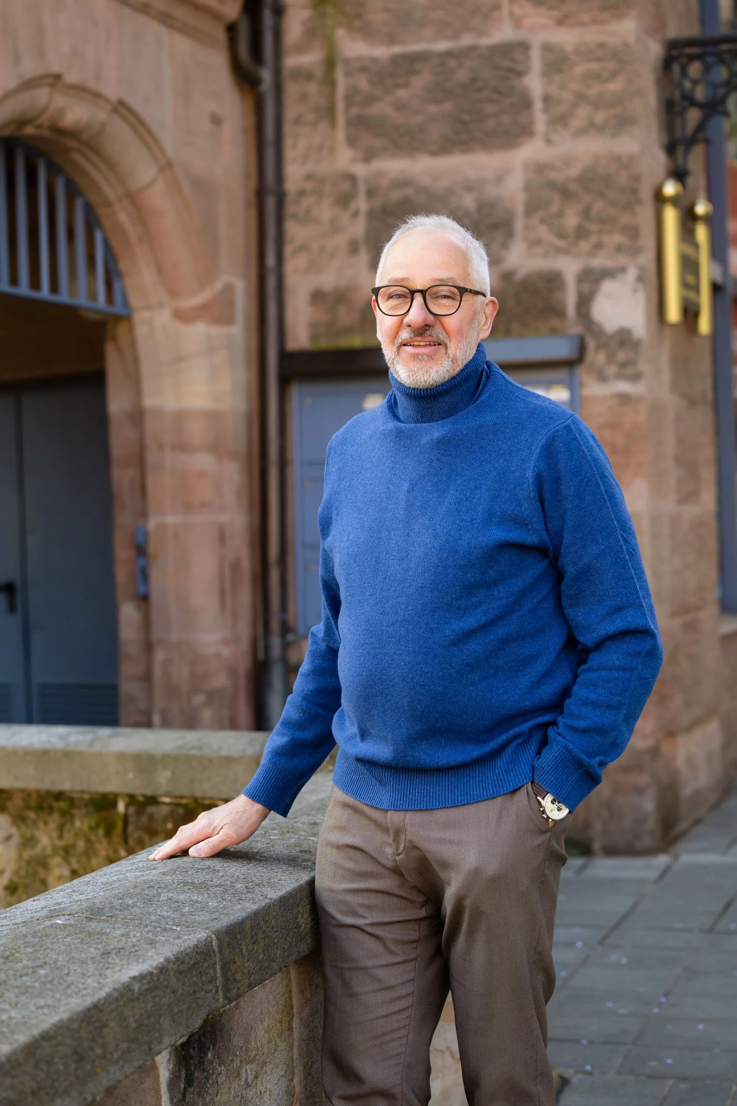 Mann mit Brille, grauem Bart, in blauem Pullover, lächelnd vor einer alten Steinmauer, in der Stadt.