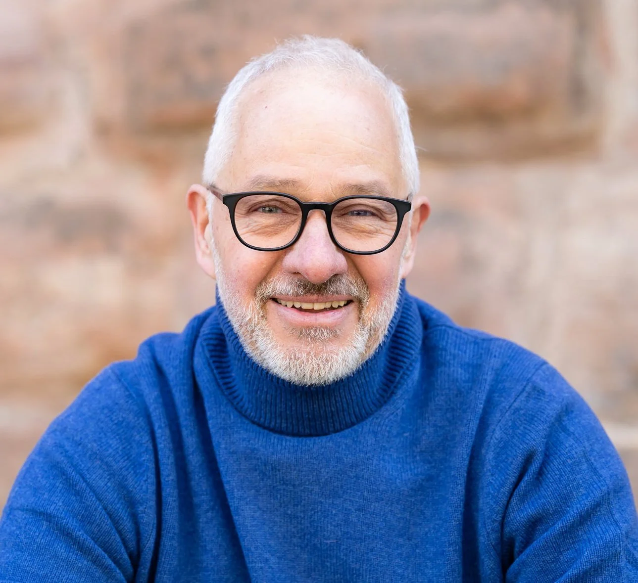Ein älterer Mann mit weißen Haaren, Brille und grau-weißem Bart trägt einen blauen Pullover und lächelt in die Kamera, im Hintergrund ein verschwommener, heller Steinwand.