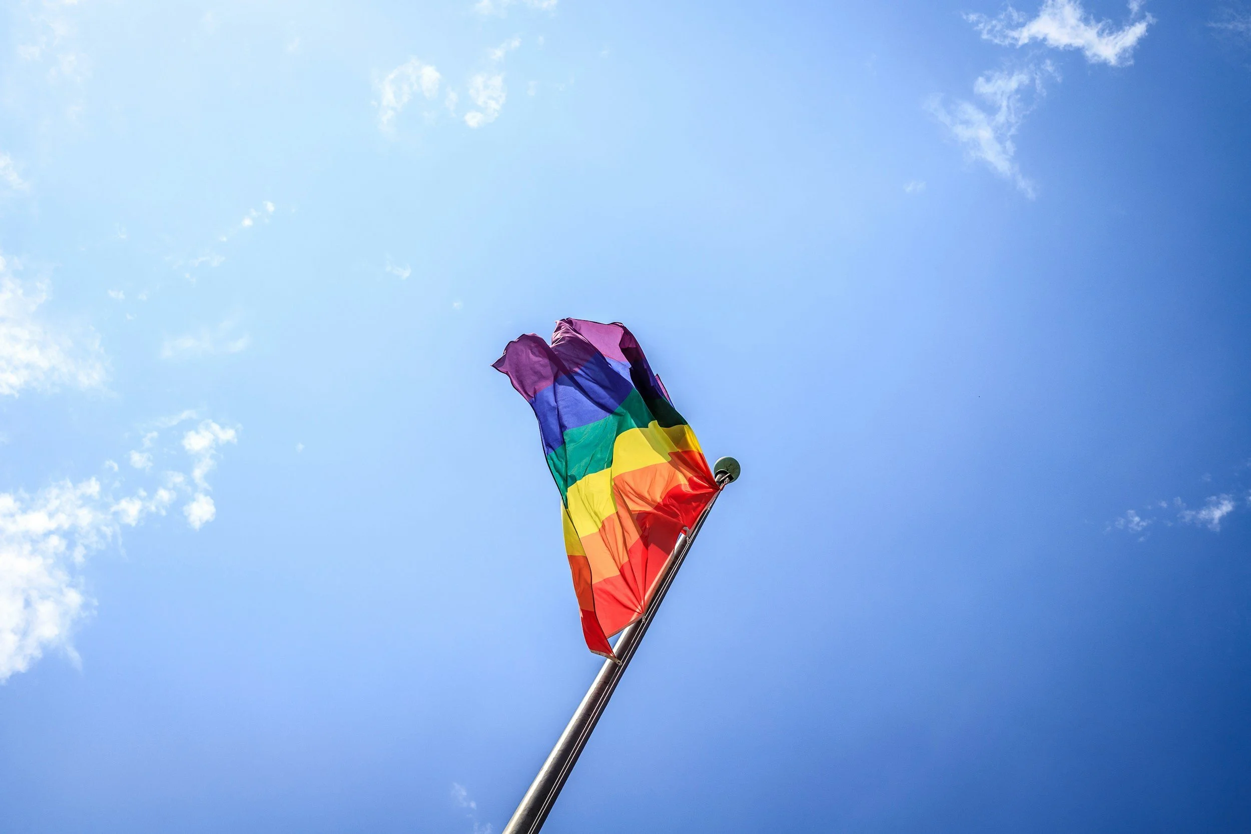 A rainbow pride flag flying on a flagpole against a clear blue sky with a few scattered clouds.