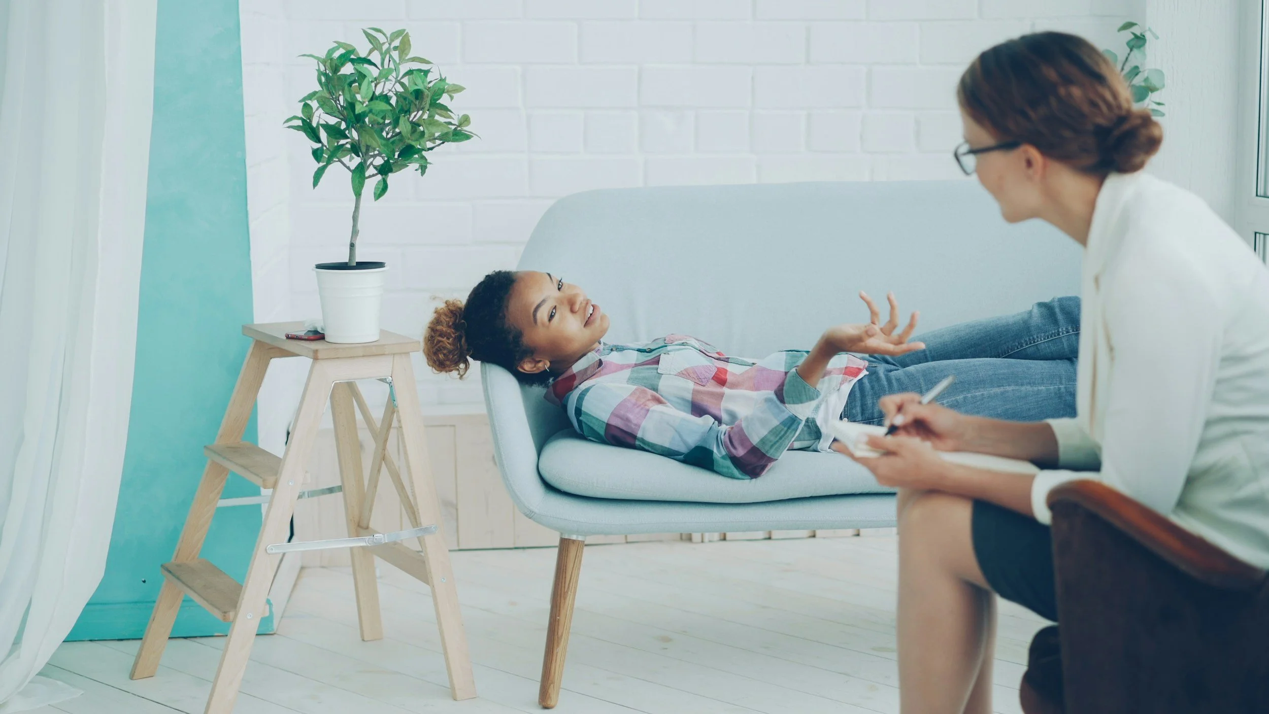 A woman sitting on a chair takes notes while a young woman lies on a light blue sofa, talking and gesturing with her hand. A potted plant on a small wooden table is in the background.