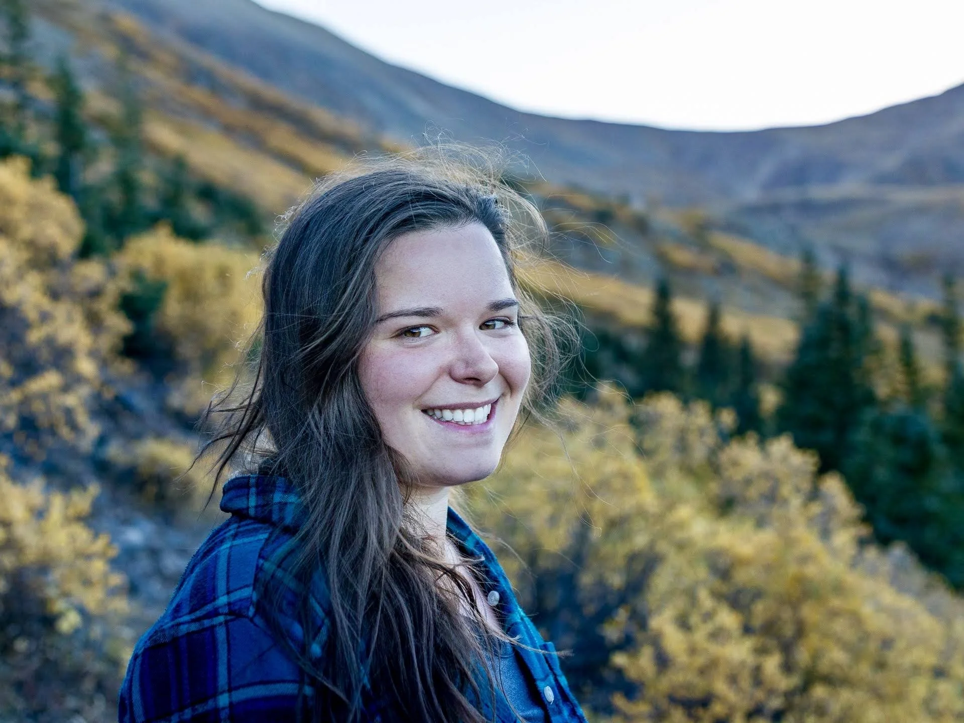 A young woman with long brown hair, smiling, outdoors with mountains and autumn trees in the background.