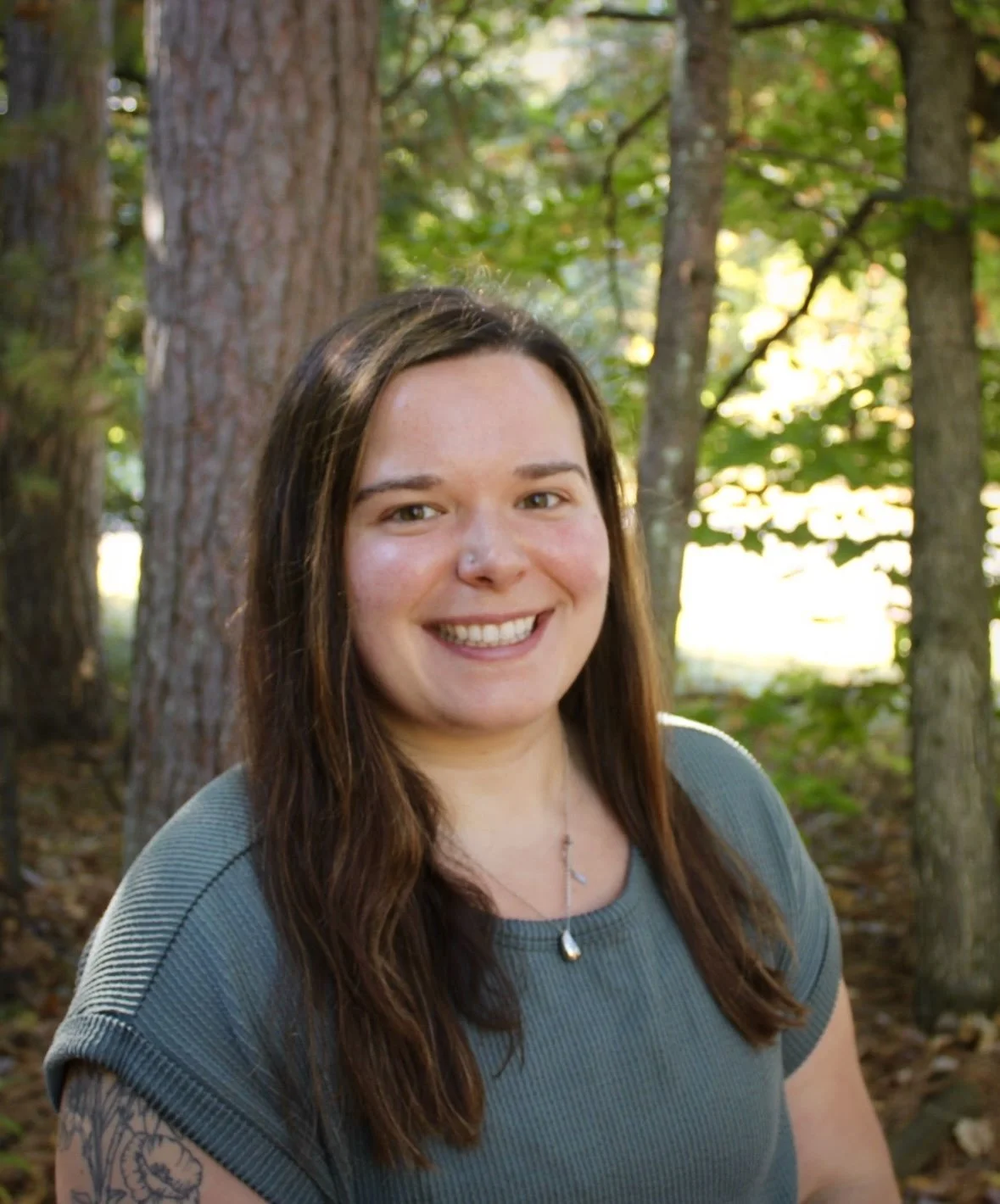 A woman with long brown hair smiling outdoors among trees in a forest setting.
