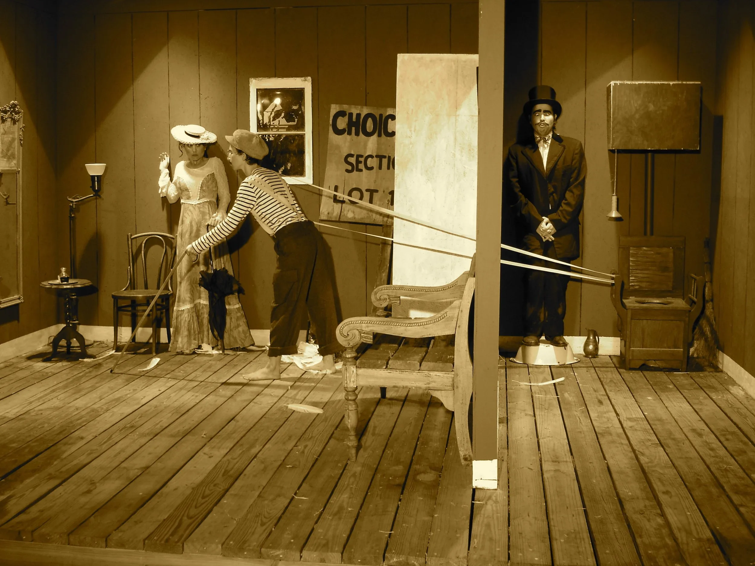 A vintage-style diorama scene with mannequins of two women and a man, one woman leaning on a chair, the other woman in a dress and hat, and the man in a suit and top hat standing in a partitioned room with wooden flooring, chairs, a small table, and props.