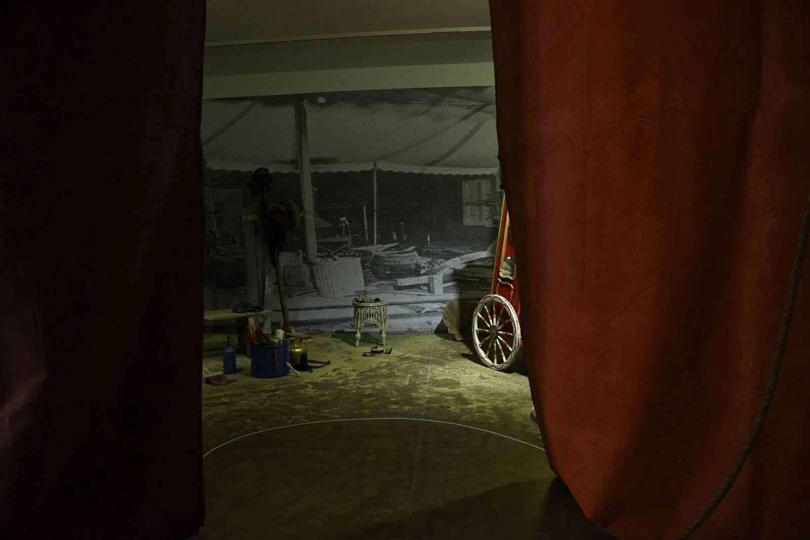 A dimly lit backstage area with red curtains partially parted, revealing a gray wall with a vintage black and white photograph. Items on the floor include a small chair, a wheel, and various tools and containers.